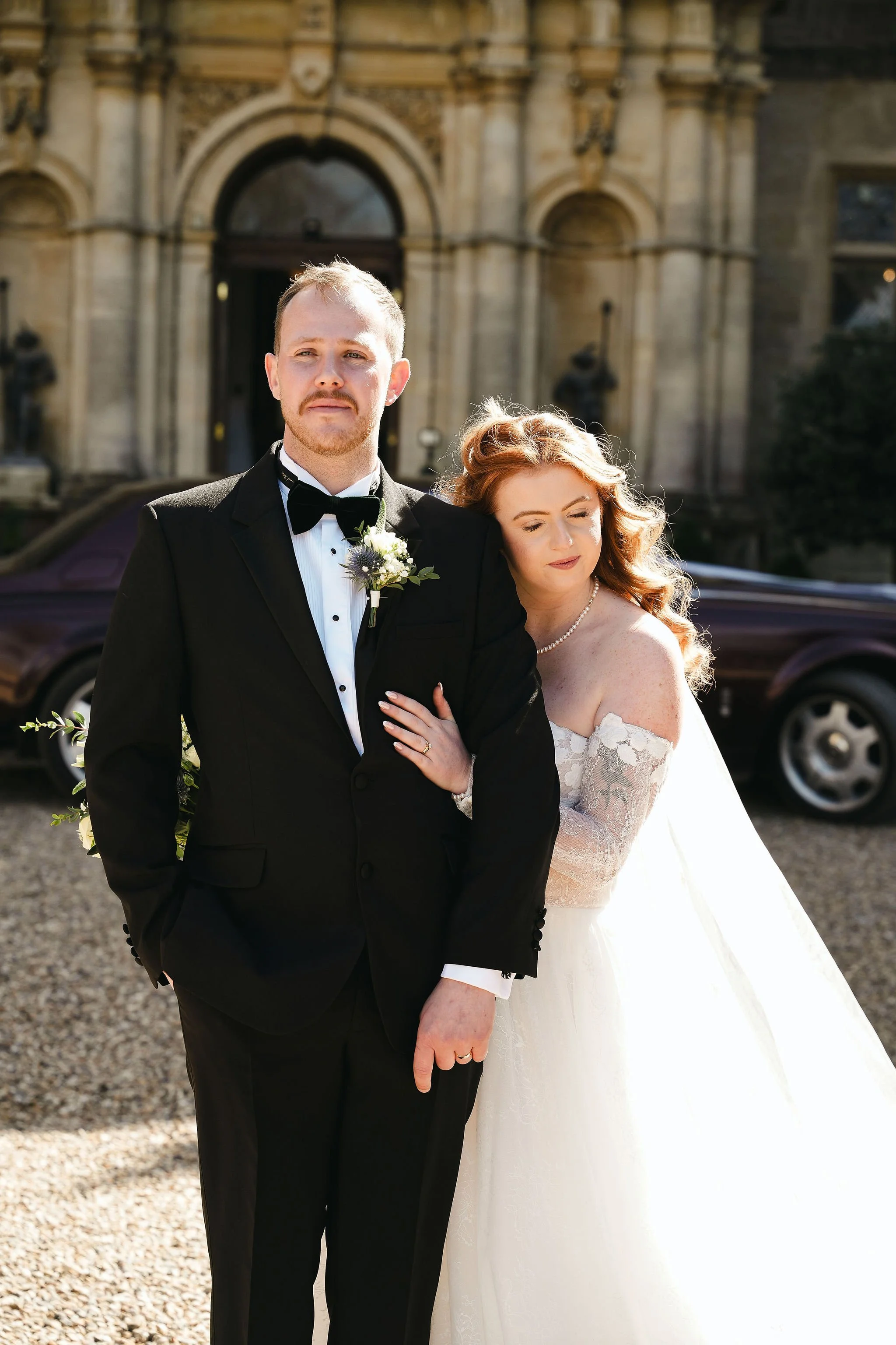 A bride and groom standing outdoors in front of a historic building, dressed in wedding attire. The groom is wearing a black tuxedo with a bow tie, and the bride is in a white wedding gown with lace details and an off-the-shoulder design. The bride h
