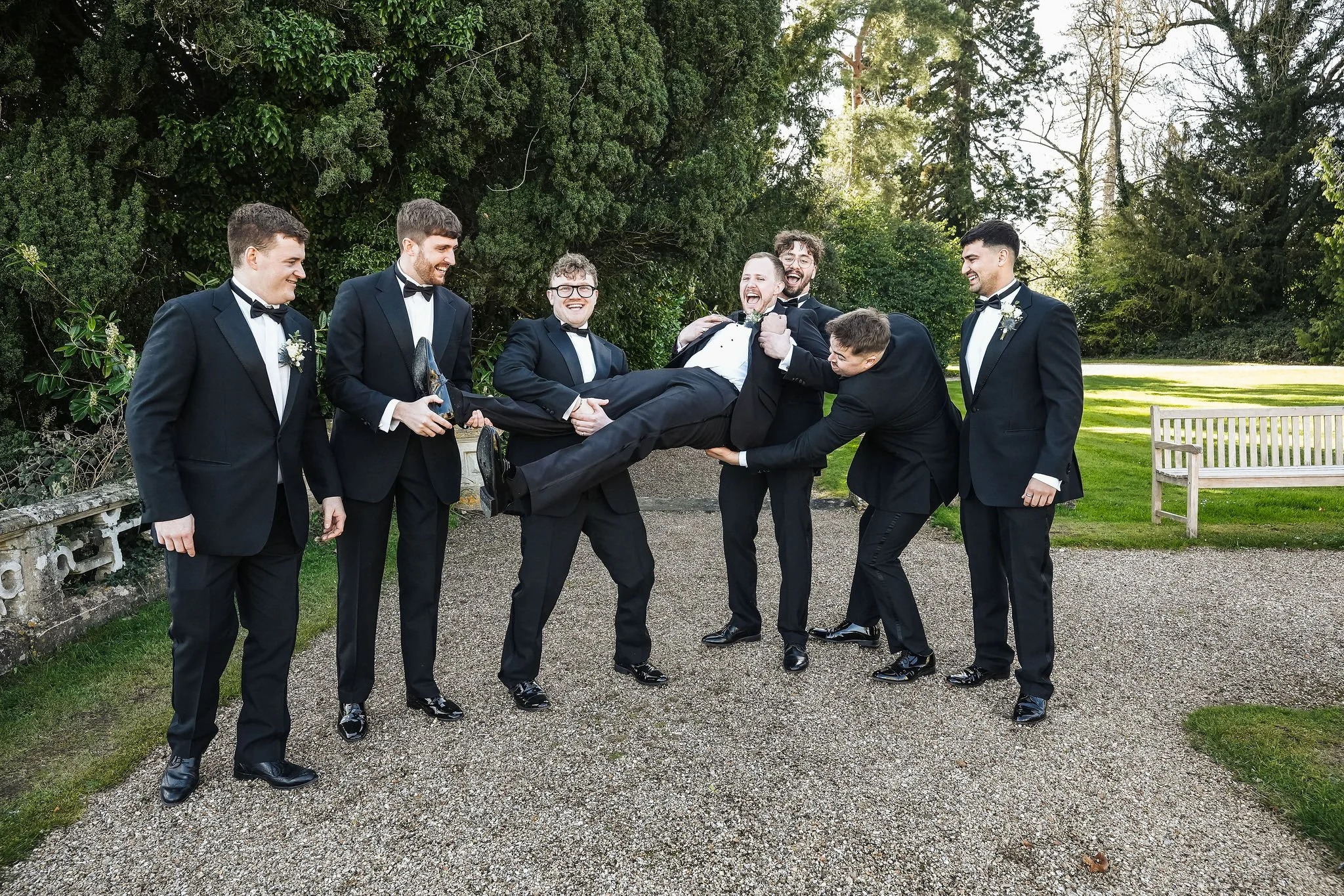 Group of seven men in black suits and bow ties, smiling and holding another man in tuxedo, who is lying across their arms, outdoors with greenery in the background.