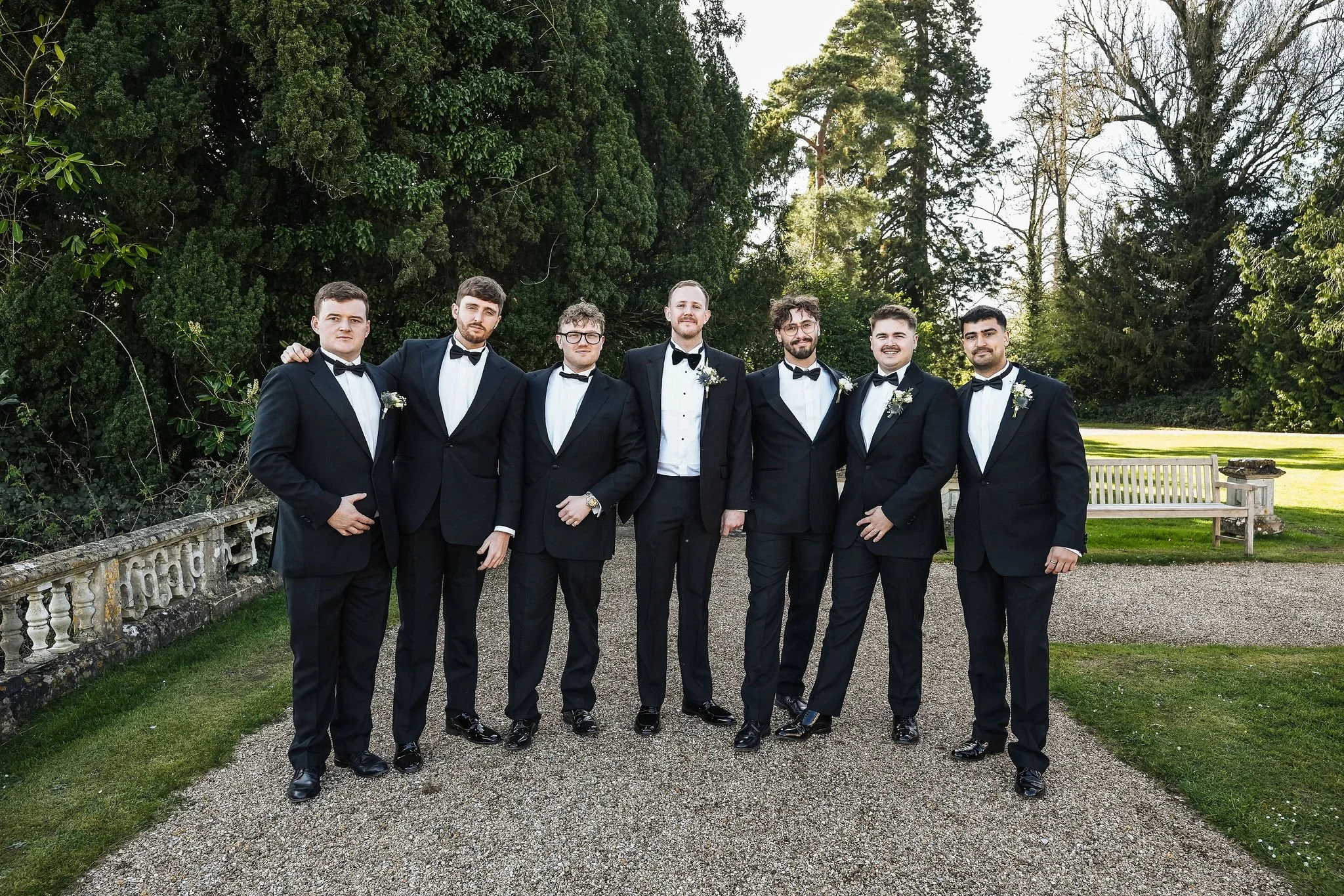 Group of seven men in tuxedos with bow ties standing outdoors on a gravel path, with green trees and a white bench in the background, posing for a photo.