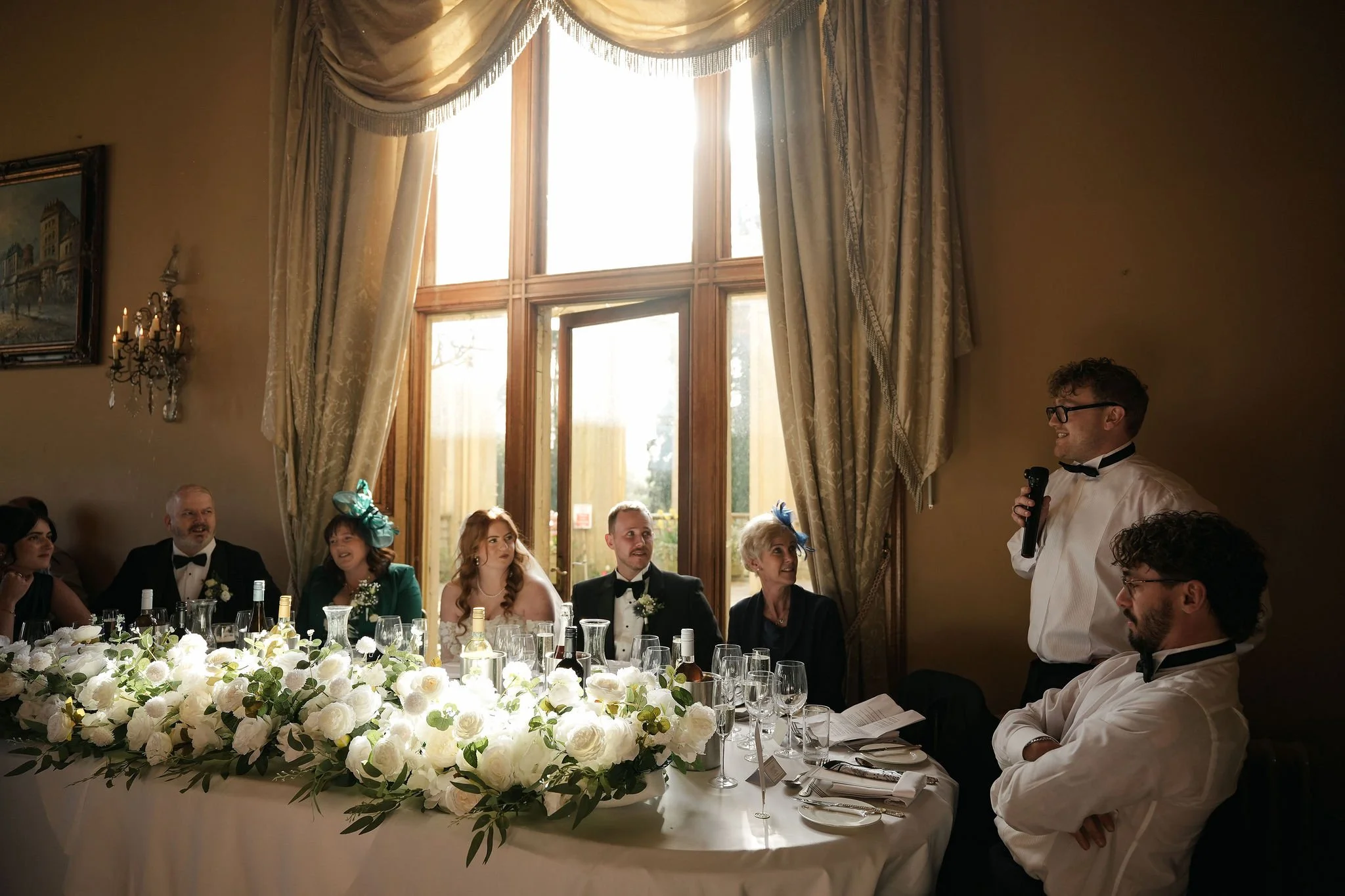 A group of people sitting at a formal table during a wedding reception, listening to a man in a white tuxedo giving a speech, with a large floral centerpiece, alcohol bottles, and glassware on the table. Large windows with beige curtains allow sunlig