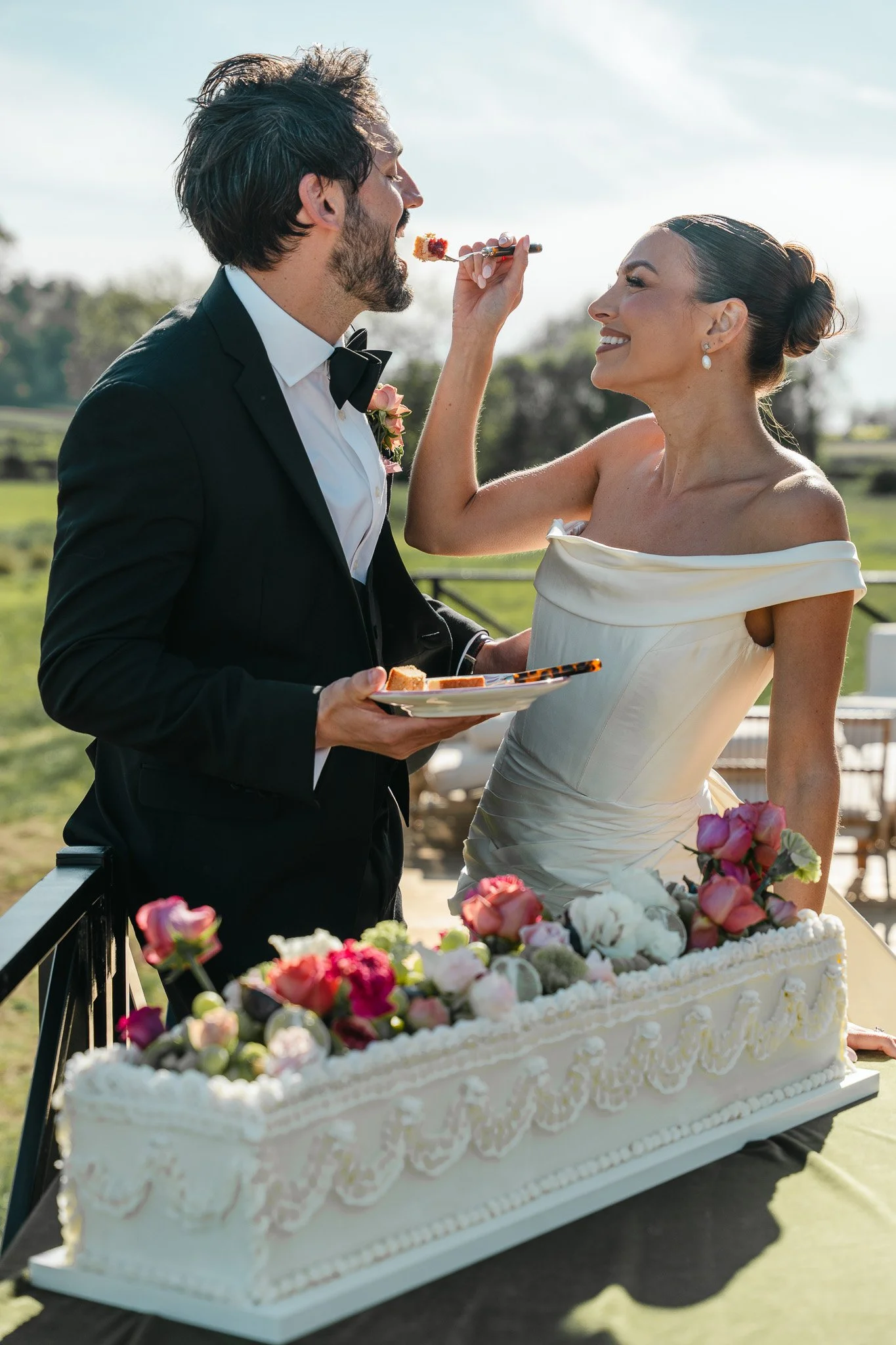 A bride and groom celebrating their wedding outdoors, with the groom feeding the bride a piece of cake, set on a decorated table with colorful flowers.
