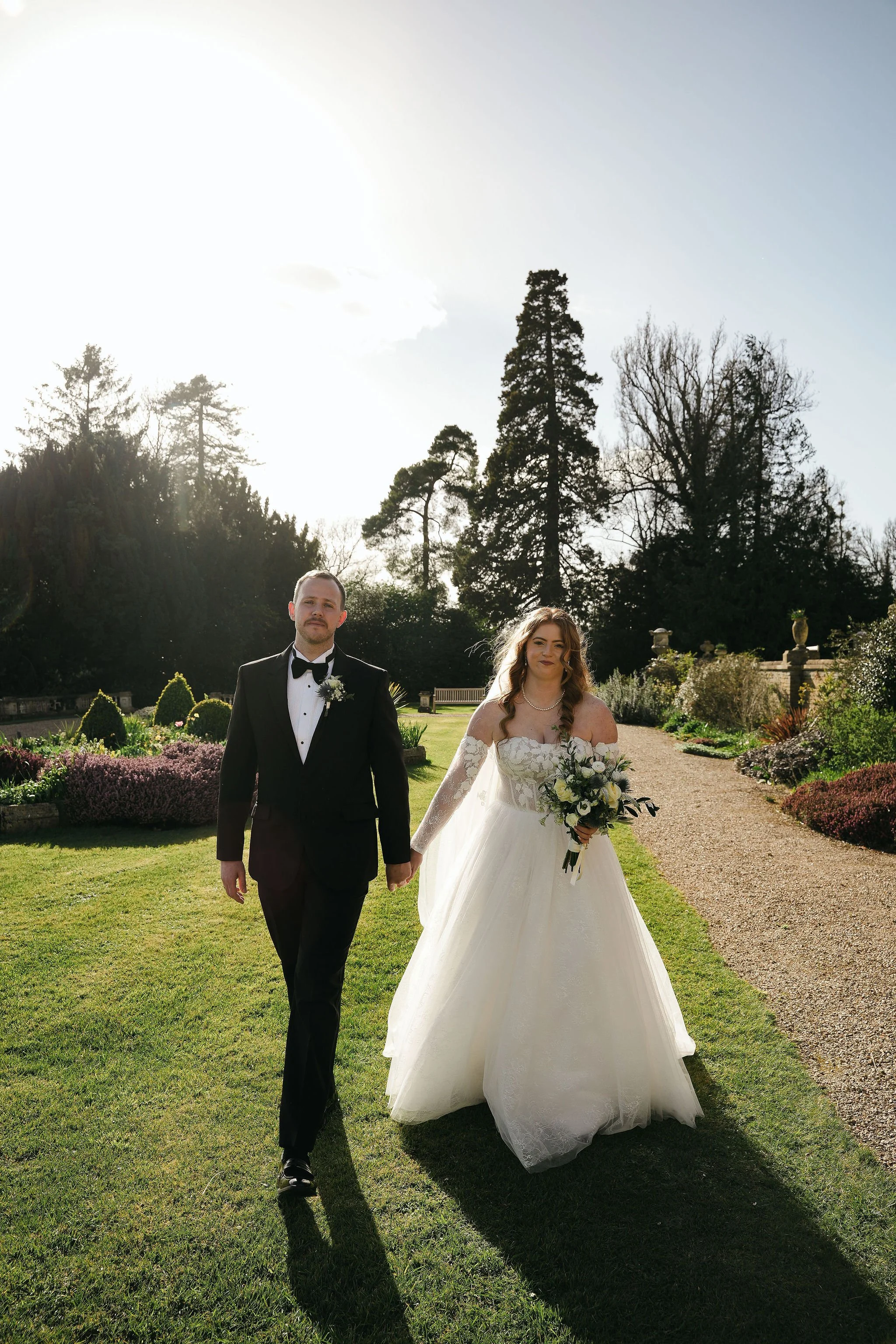 A bride and groom walking hand in hand in a garden on their wedding day. The groom is wearing a black tuxedo with a bow tie, and the bride is in a white wedding dress holding a bouquet of flowers.