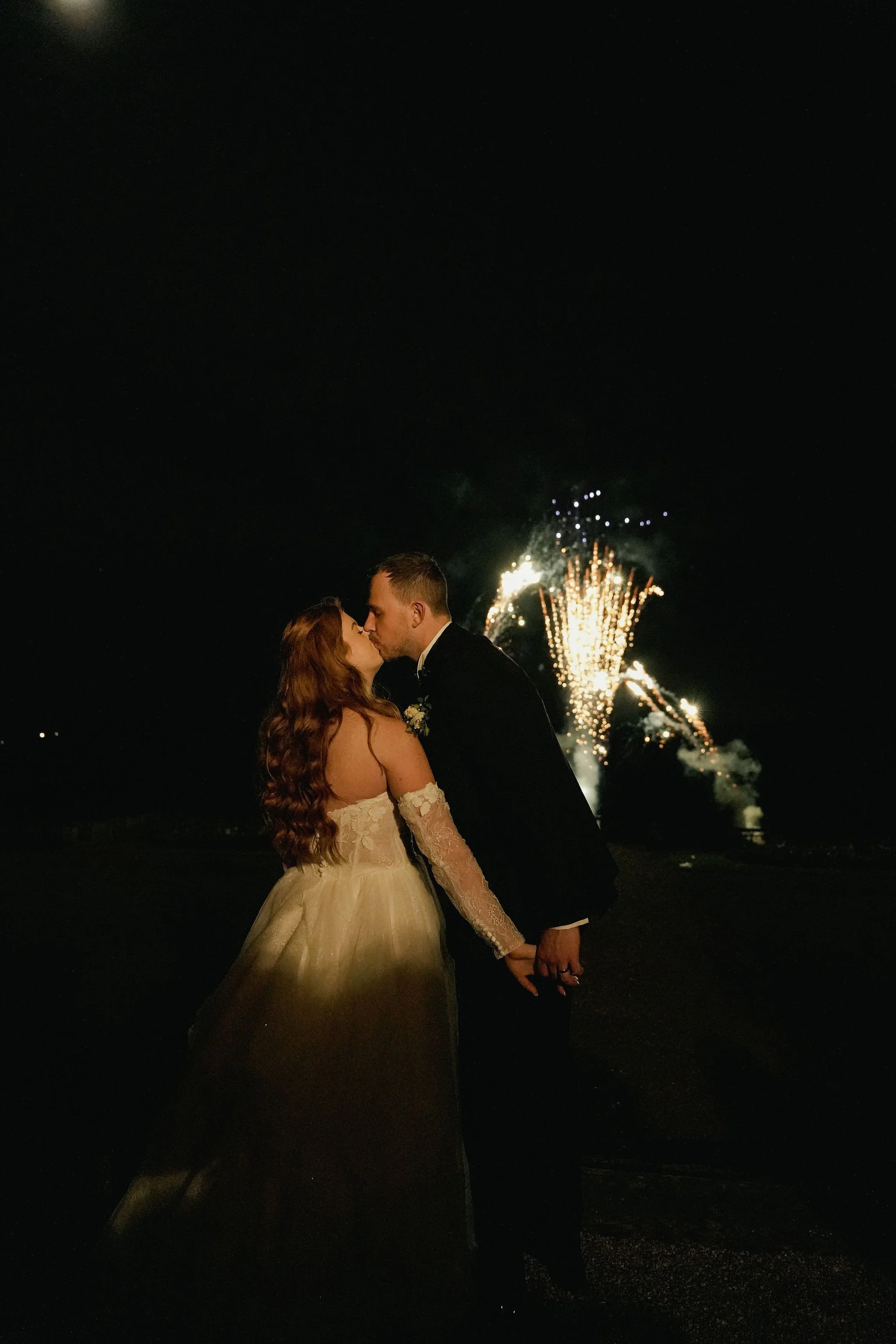 A bride and groom sharing a kiss at night, fireworks in the background.