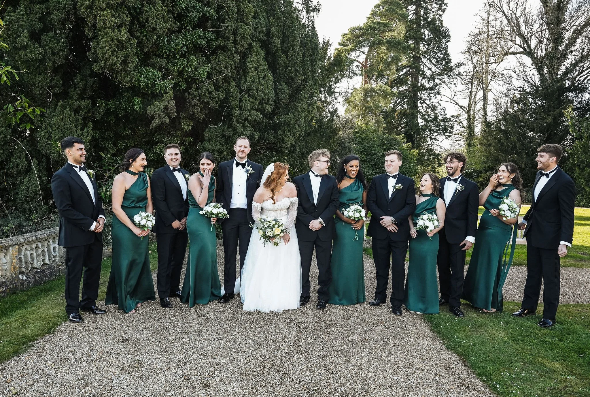 A wedding party in formal attire standing outdoors with large trees in the background. The bride is wearing a white gown, and the groom is in a black tuxedo. The bridesmaids are in green dresses, and the groomsmen are in black tuxedos. Everyone is sm