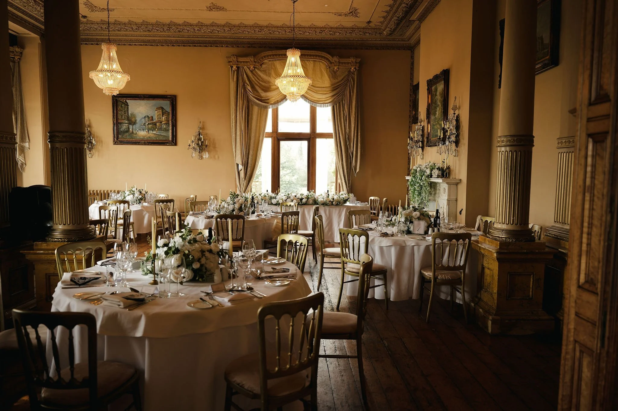 Elegant dining room decorated for a formal event with round tables covered in white tablecloths, floral centerpieces, and multiple wine glasses; tall windows with drapes and chandeliers providing warm lighting; framed landscape paintings and ornate w