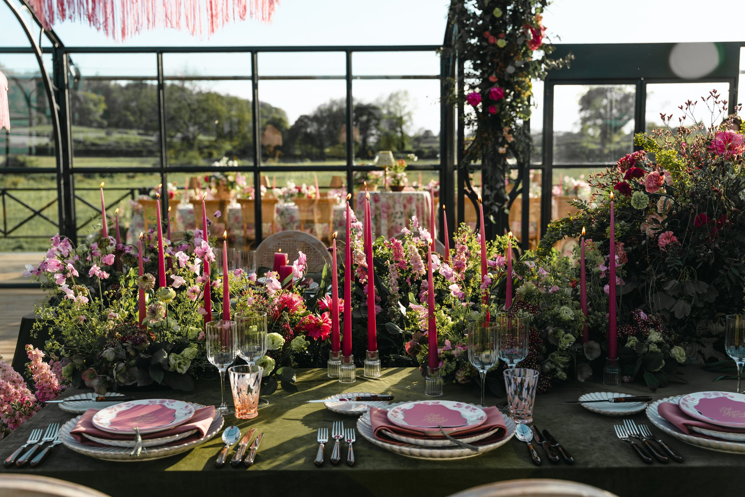 A decorated outdoor dining table with pink candles, pink plates, and glassware, surrounded by pink and purple flowers and greenery, set in a garden or vineyard area.