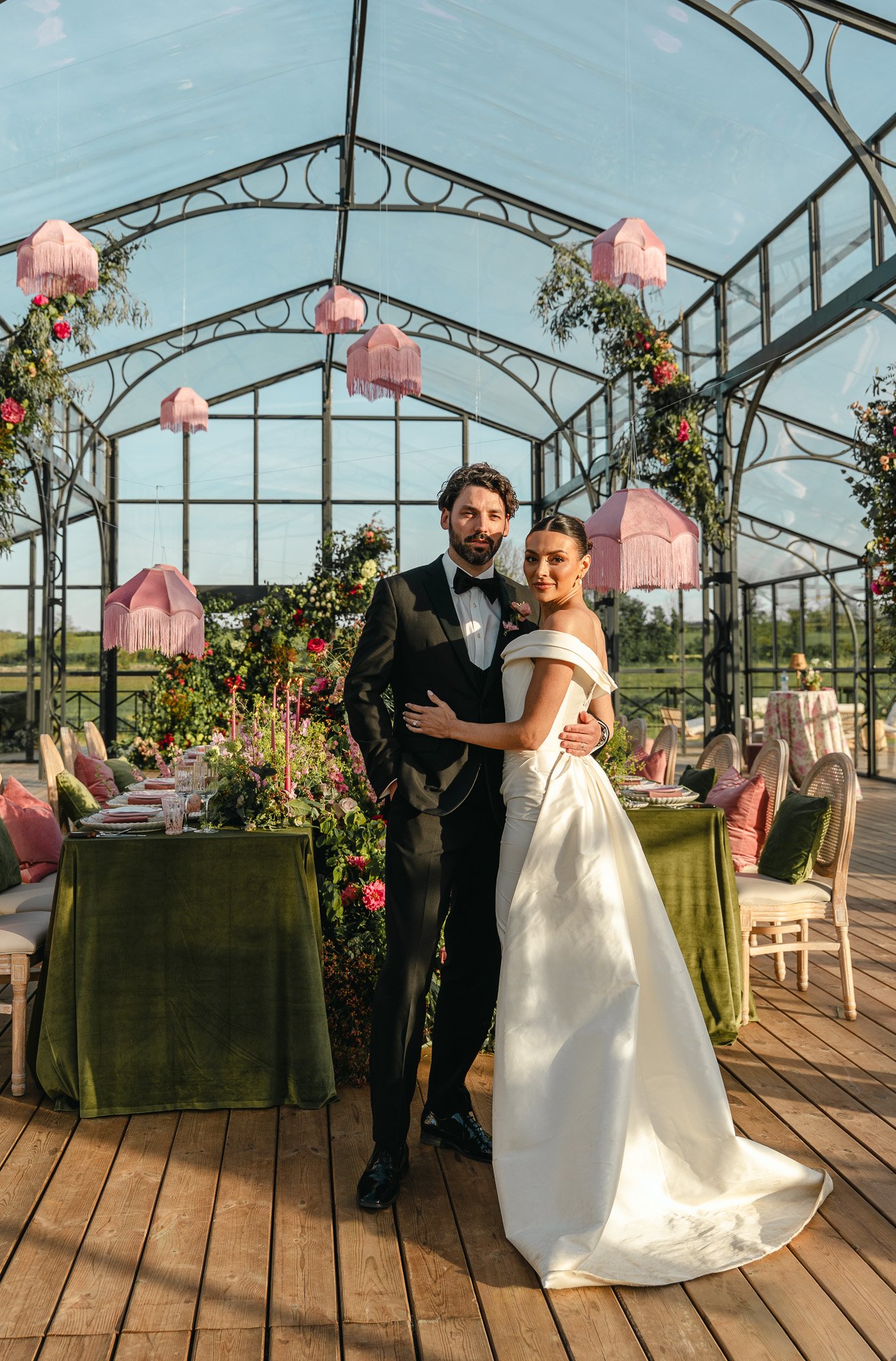 A bride and groom standing together in a decorated greenhouse during their wedding reception, with pink umbrellas hanging from the ceiling and floral arrangements on tables.