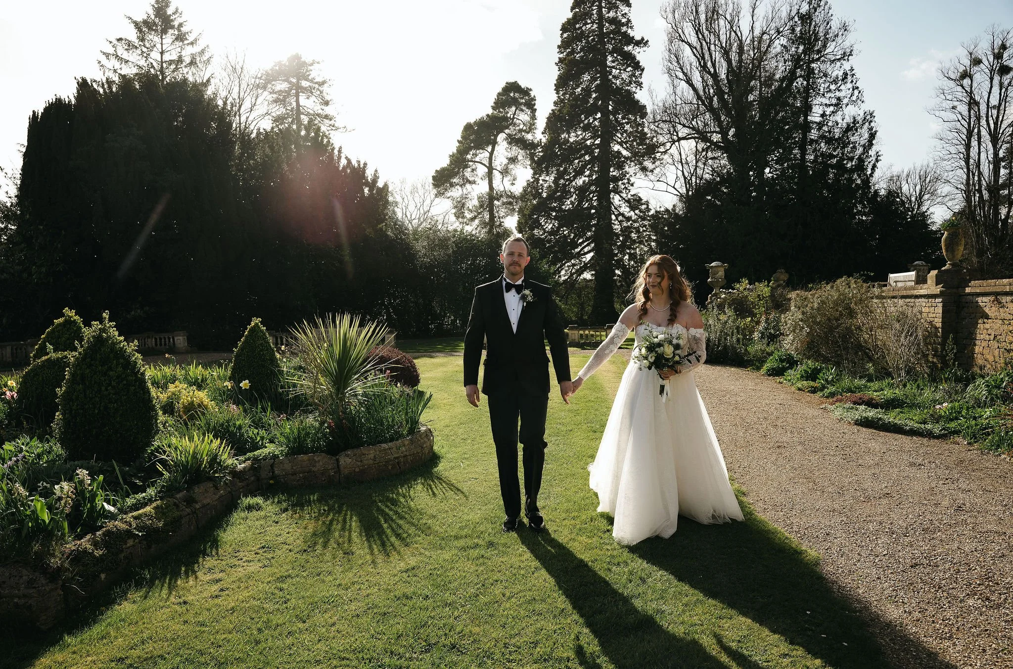A bride and groom walking hand in hand in a garden during a wedding, with tall trees and sunlight in the background.