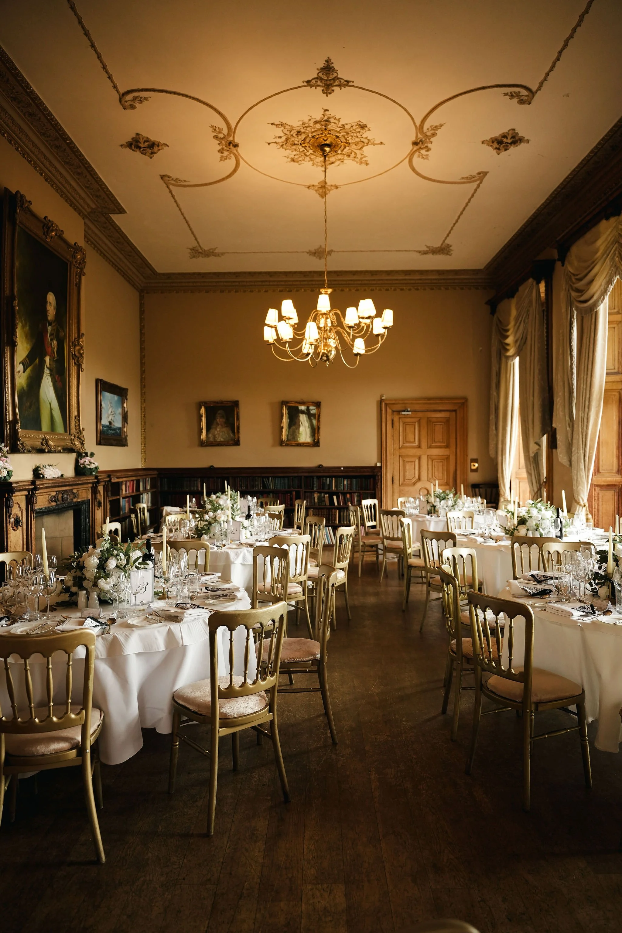 Elegant dining room set up with round tables covered in white tablecloths, decorated with floral centerpieces, surrounded by gold chairs, ornate chandelier, and classic portraits on the walls, with large windows draped in curtains.