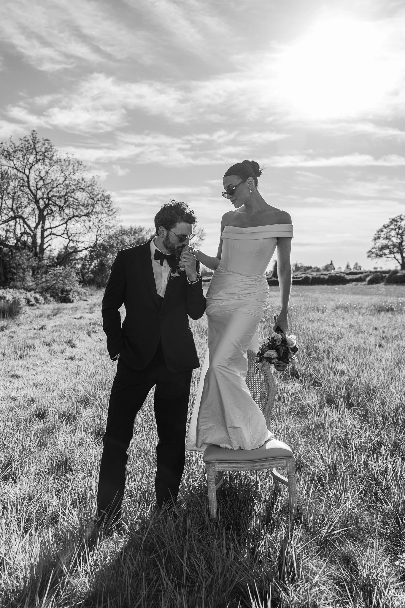 A man kneeling and kissing the hand of a woman standing on a chair outdoors. The woman is wearing a wedding dress, and the man is in a tuxedo with glasses. The scene is set in a grassy field with trees and cloudy sky in the background.