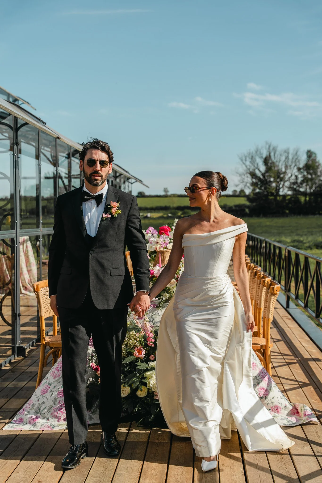 A bride and groom holding hands and walking on a decorated outdoor wedding altar with flowers and floral tablecloths, under a clear blue sky, on a wooden deck with a grassy field in the background.