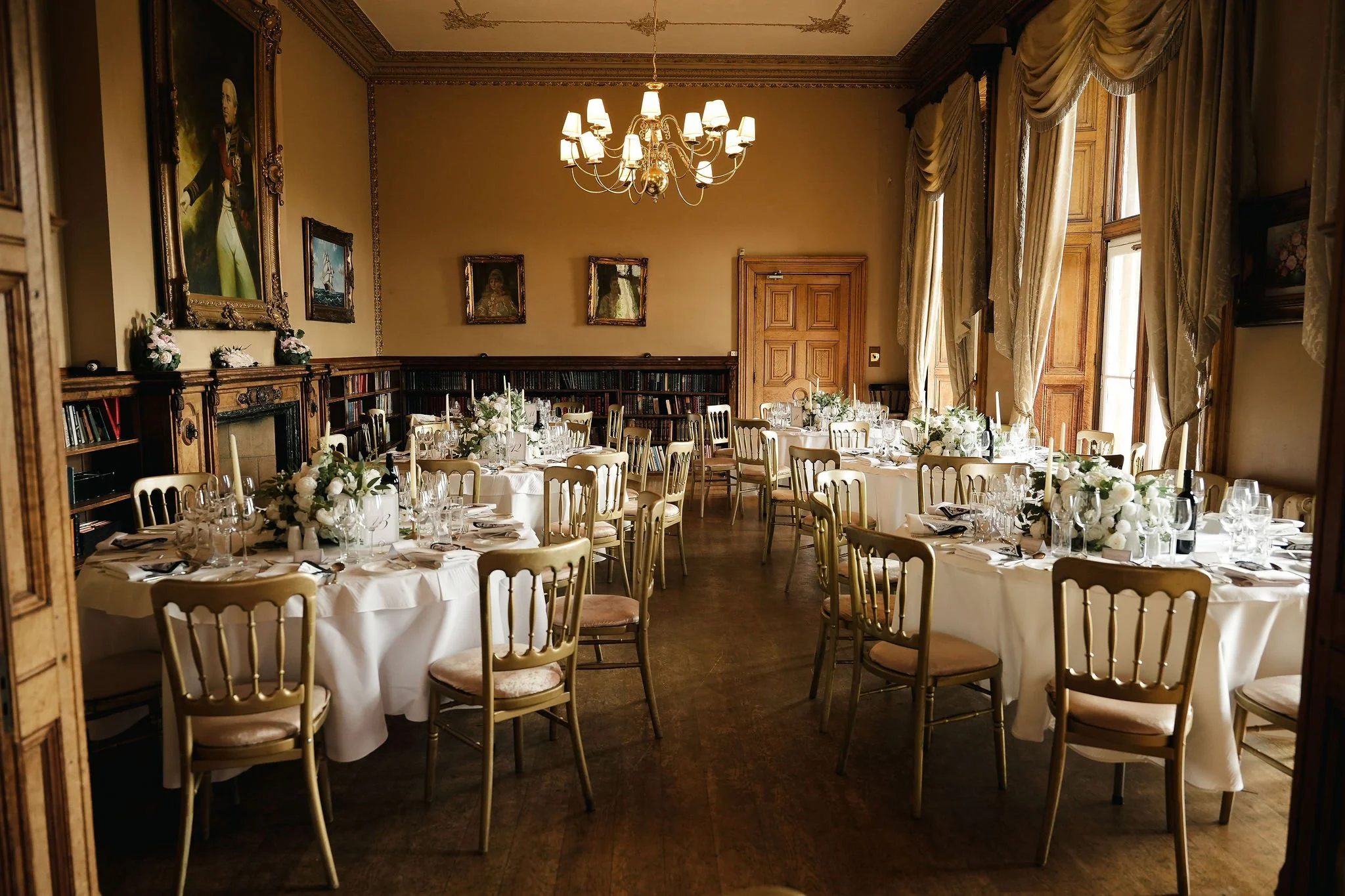 Elegant dining room with round tables covered in white tablecloths, decorated with white floral centerpieces, arranged for a formal event or wedding reception. The room features wooden paneled walls, ornate curtains, and a chandelier hanging from the