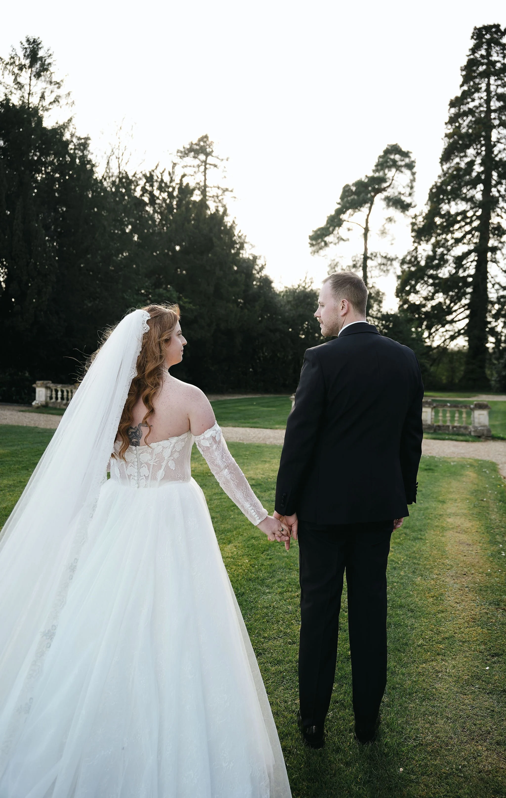 A bride and groom holding hands and walking outdoors on a grassy area with trees in the background. The bride is wearing a white lace wedding dress with off-the-shoulder sleeves and a long veil, while the groom is dressed in a black suit. They are lo