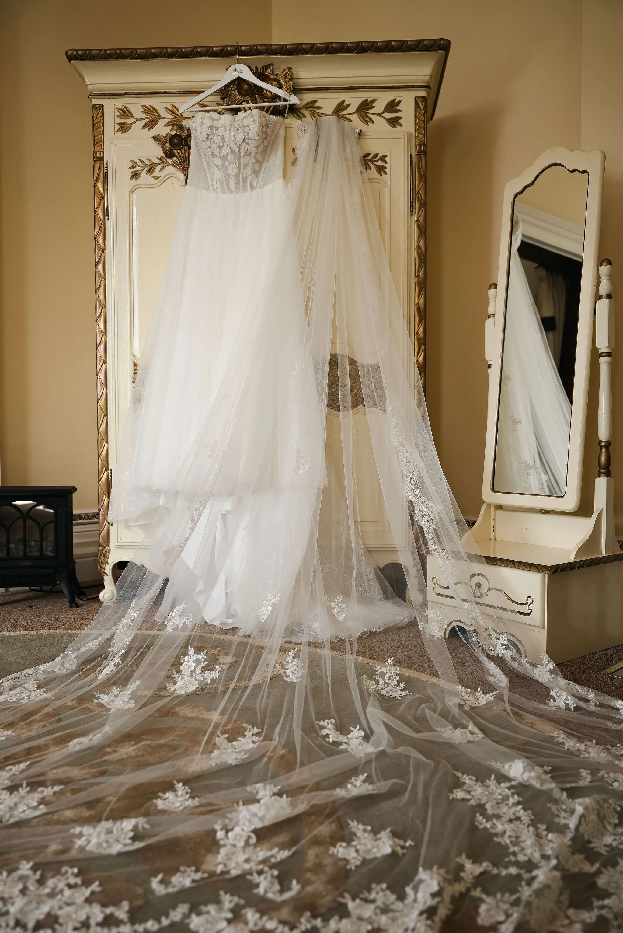 A wedding dress with lace and tulle hangs on a wooden closet door, with a long embroidered veil flowing onto the floor in front of it. A mirror reflects part of the dress and vanity.