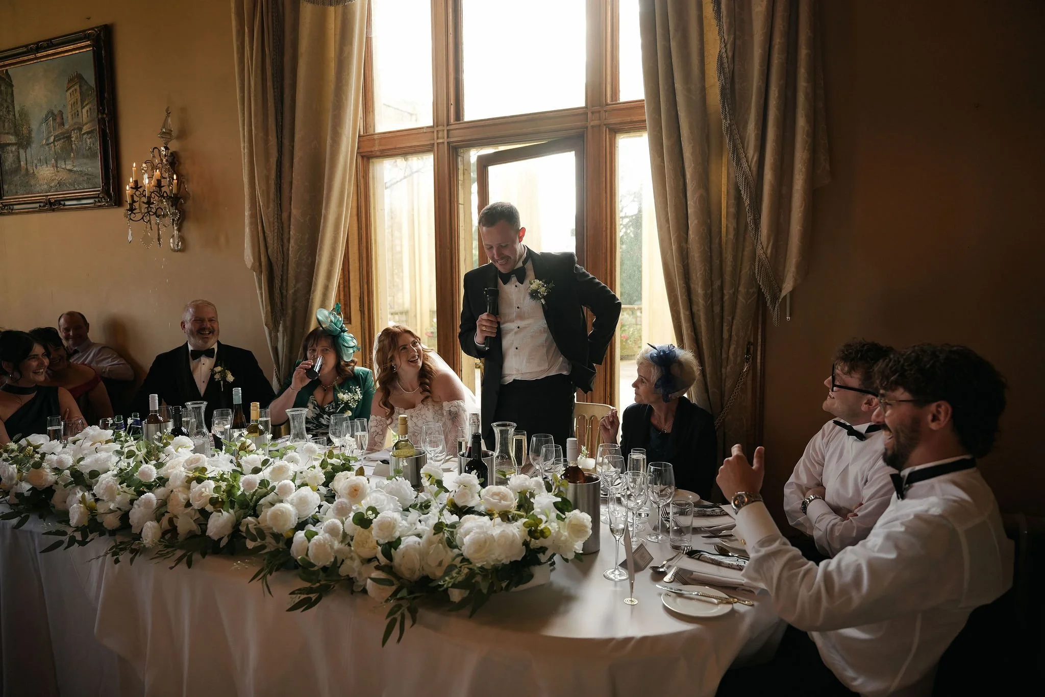 A group of people sitting at a wedding reception table, with a decorated centerpiece of white flowers, enjoying a speech by a man in a tuxedo holding a microphone, in a room with large windows and cream-colored curtains.