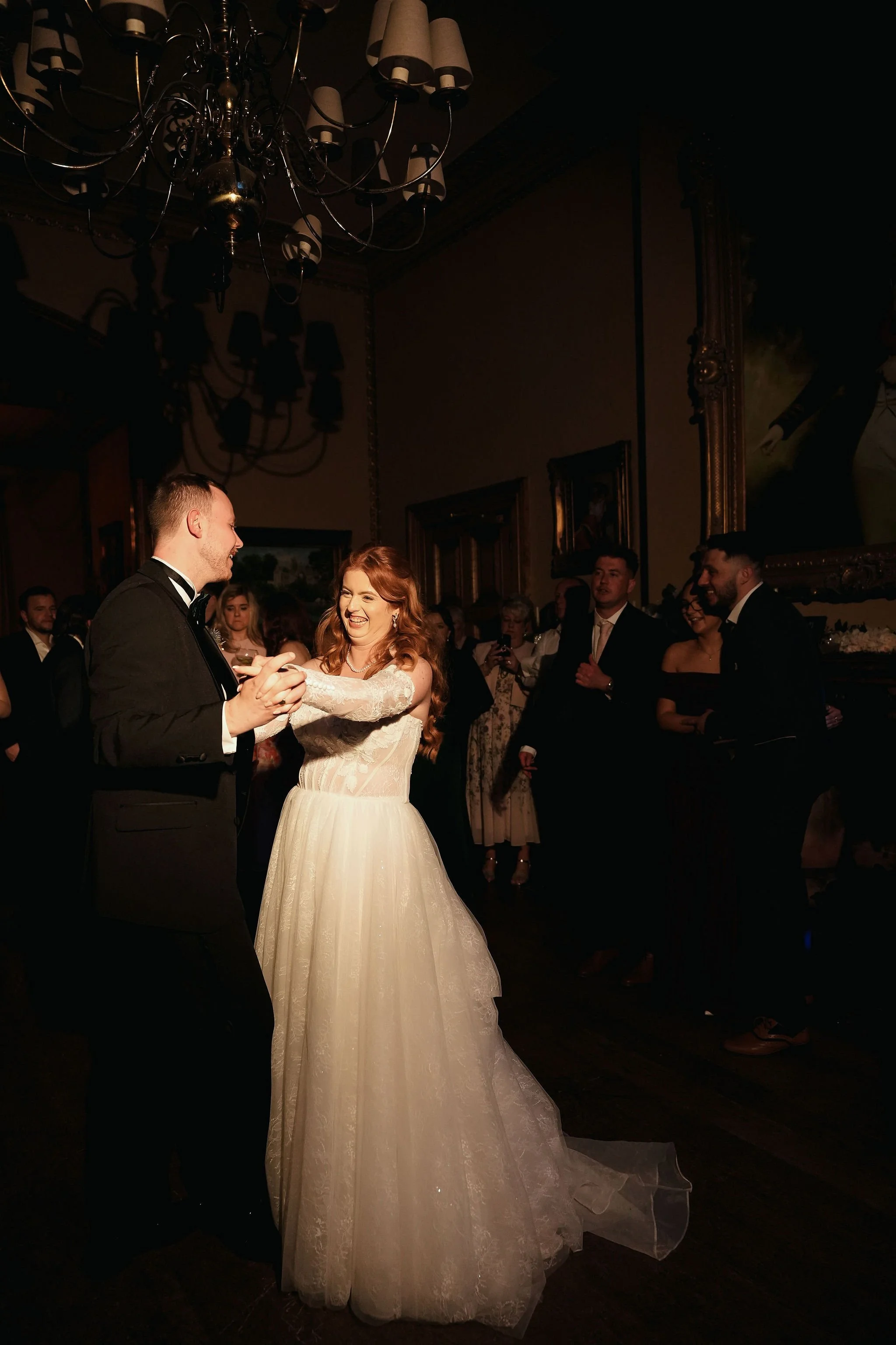 A bride and groom dancing at their wedding reception, surrounded by guests. The bride is smiling and wearing a white wedding gown, while the groom is in a black tuxedo. The room has dark walls, ornate frames, a large chandelier, and dim lighting.