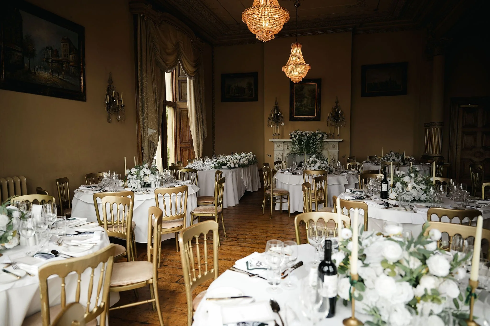 Elegant banquet room with large round tables covered in white tablecloths, decorated with white floral centerpieces and candle holders, gold chairs, and set with glassware and utensils. The room features tall windows with heavy drapes, framed paintin