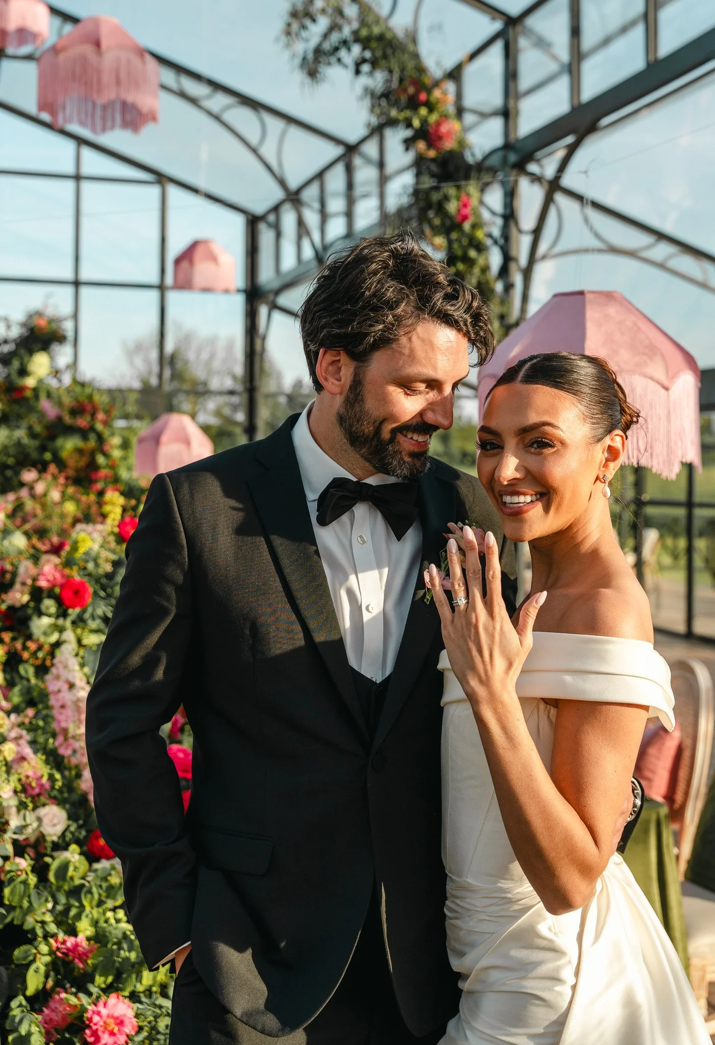 A newlywed couple stands close together outdoors, smiling and showing off a wedding ring, with Love in Bloom backyard garden setting and pink floral decorations behind them.