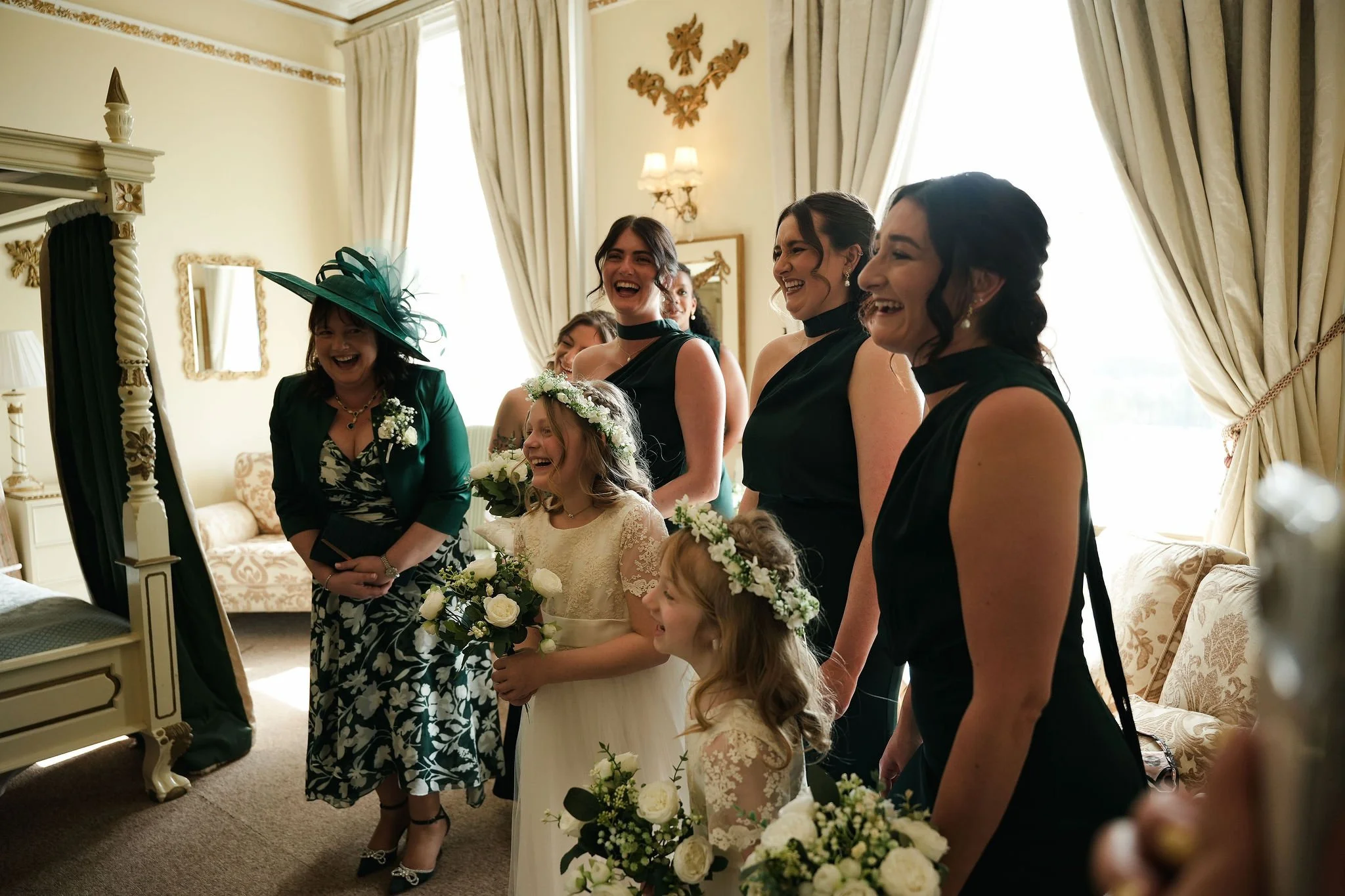 A group of women and young girls, dressed in formal attire, are laughing and smiling in a well-lit, elegant room. Some women are wearing black dresses, and the girls are in white dresses with floral wreaths.
