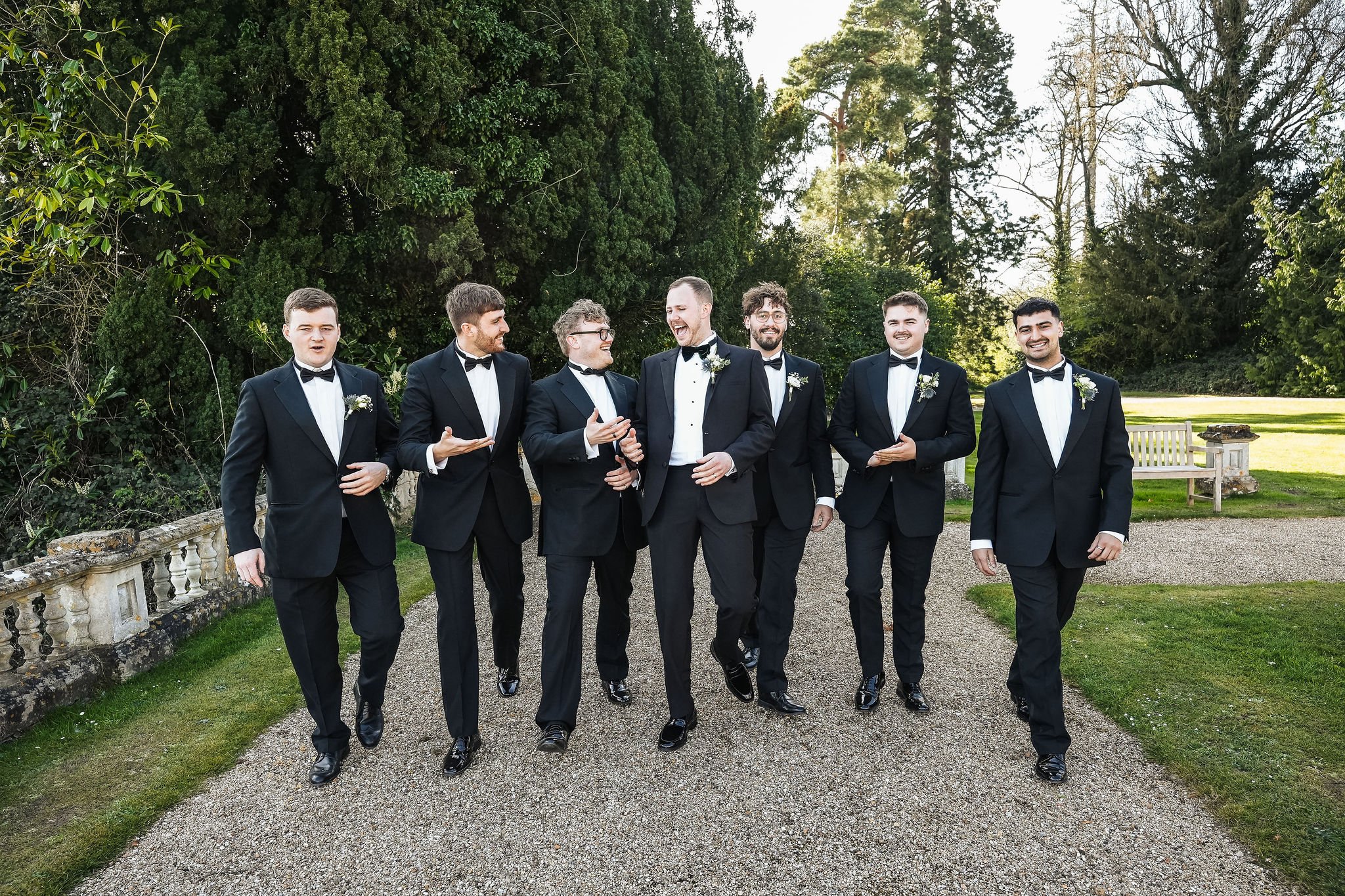 Group of seven men dressed in black tuxedos with bow ties, walking outdoors on a gravel pathway with green trees in the background, some men are smiling and laughing.