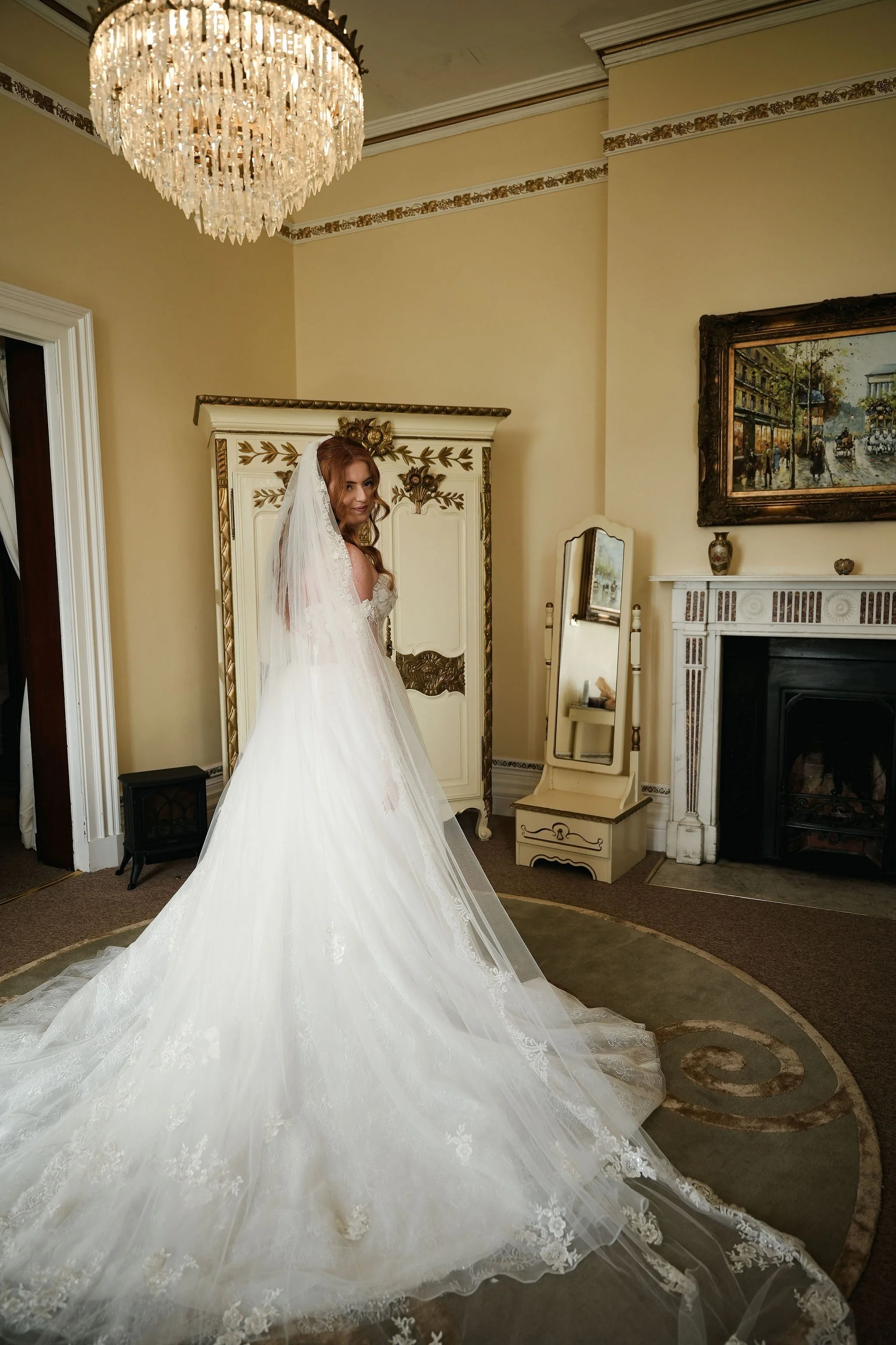 A bride in a white wedding gown with a long train and veil standing in a room with ornate decor, a chandelier overhead, a fireplace, and a painting on the wall.