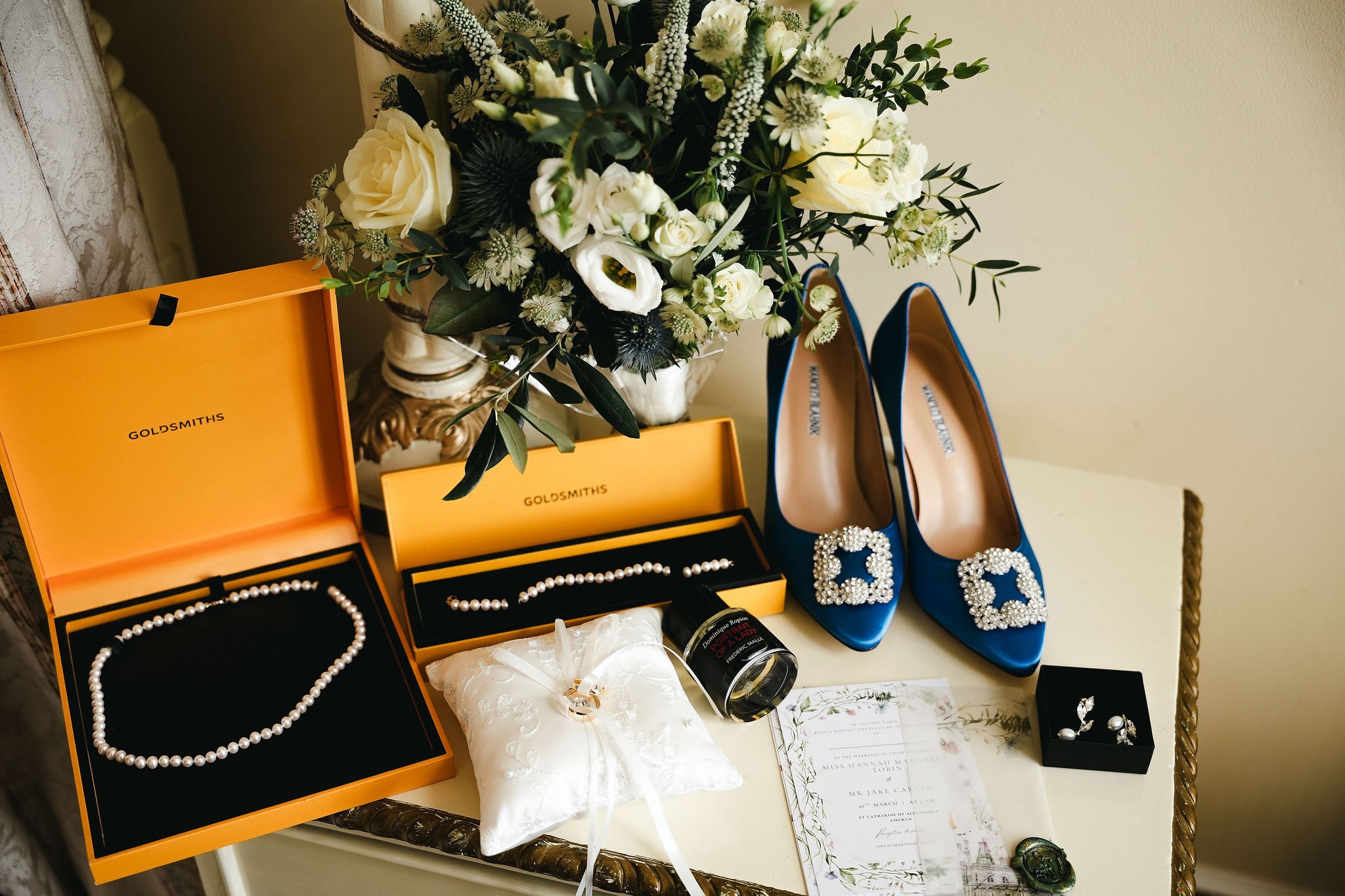 A collection of jewelry, including pearl necklaces and earrings, along with a bouquet of white flowers, a pair of blue high-heeled shoes with jeweled embellishments, and wedding invitations, all arranged on a surface for a special occasion.