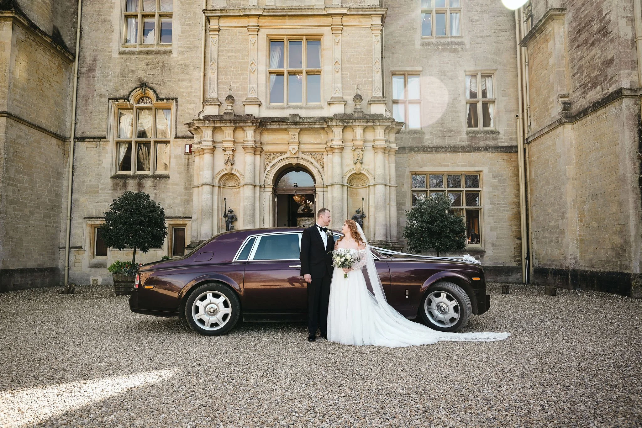 A bride and groom standing next to a vintage dark purple Rolls Royce car in front of a historic stone mansion. The bride is holding a bouquet and wearing a white gown with a long train, and the groom is in a black tuxedo. The mansion has large window
