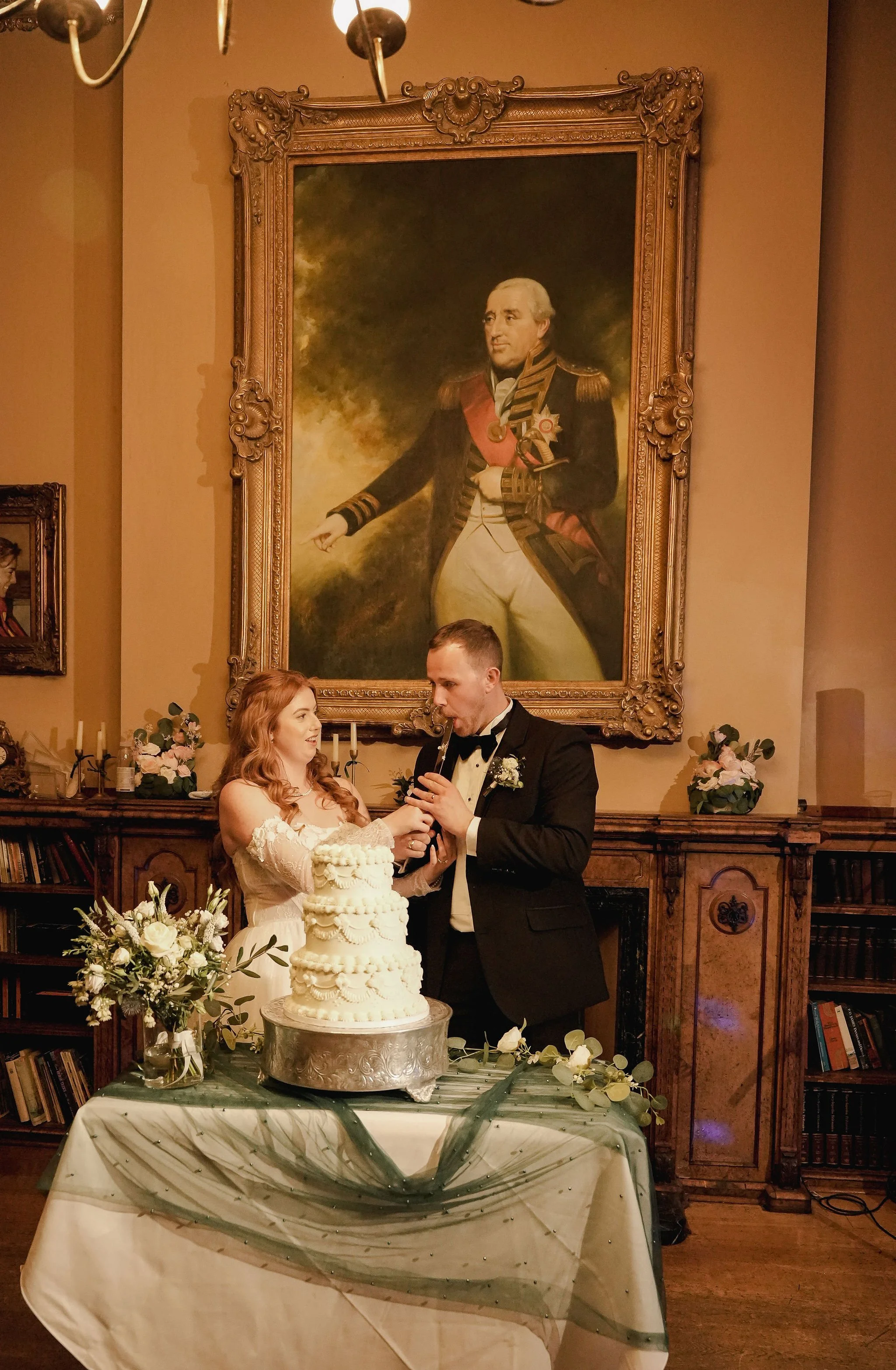 A bride and groom at their wedding, standing in front of a decorated wedding cake with flowers and greenery. In the background, there is a large, framed portrait of a historical military figure in uniform.