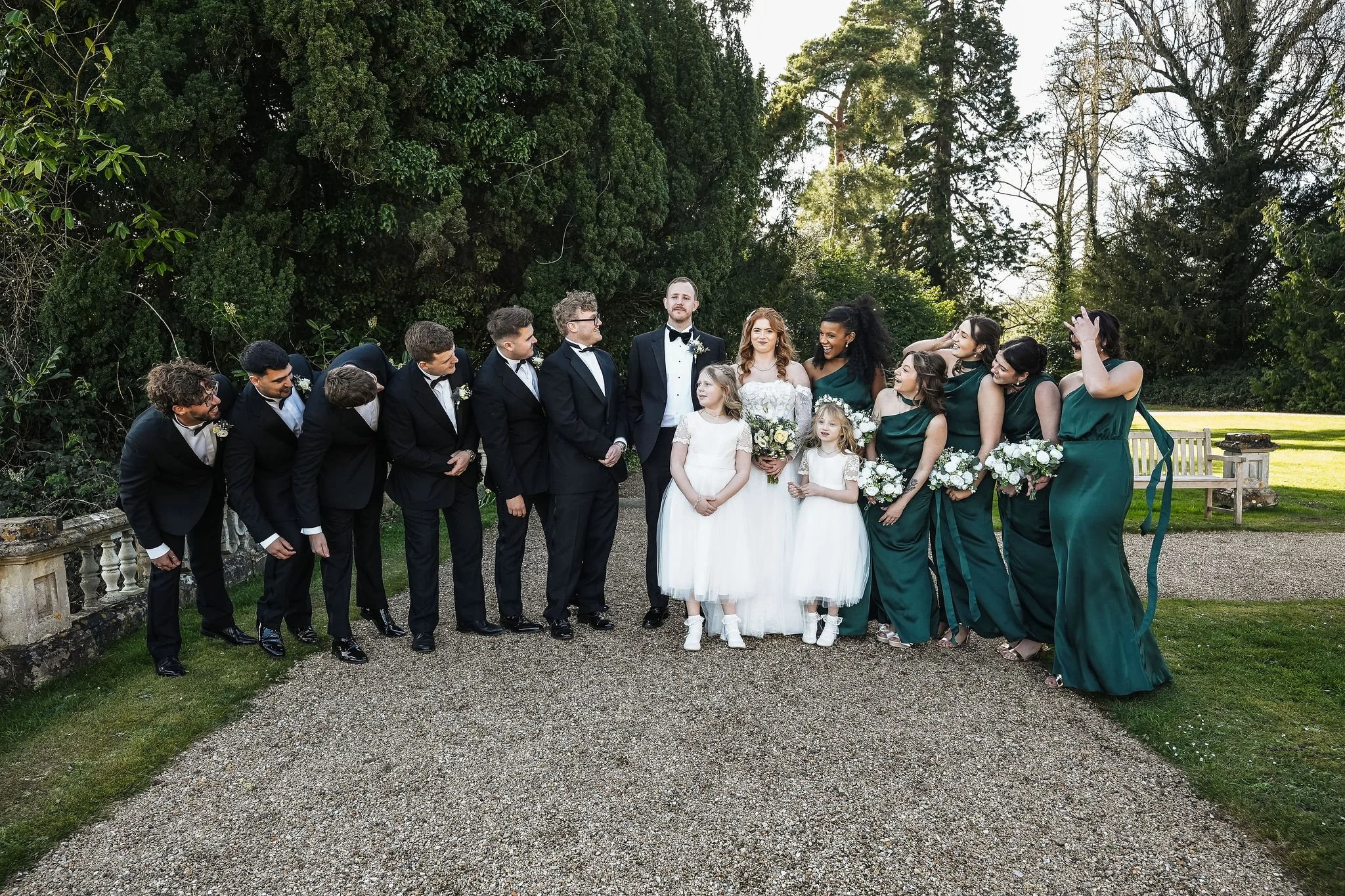Group of wedding guests, including men in tuxedos and women in green dresses, standing outdoors in front of tall trees and greenery.