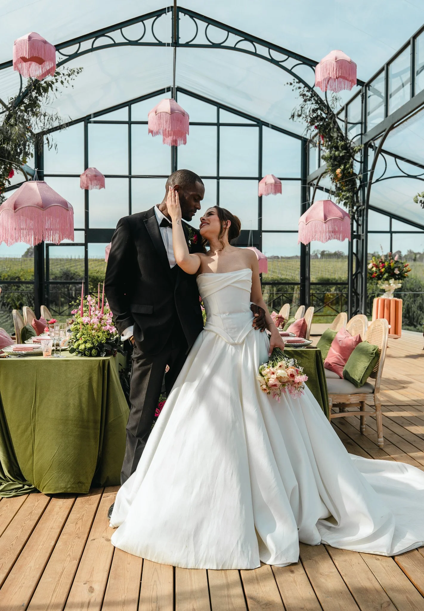 A bride and groom sharing an intimate moment during their wedding reception in a glass greenhouse with pink hanging lamps and decorated tables.