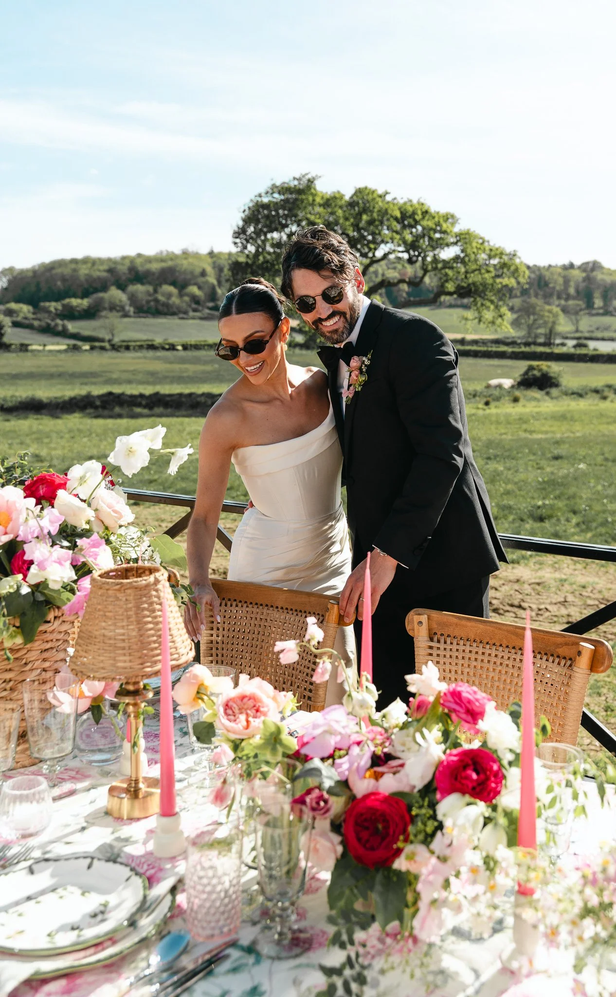A newlywed couple laughing at a wedding reception outdoors, surrounded by a decorated table with flowers and candles, in a scenic grassy landscape with trees and blue sky.