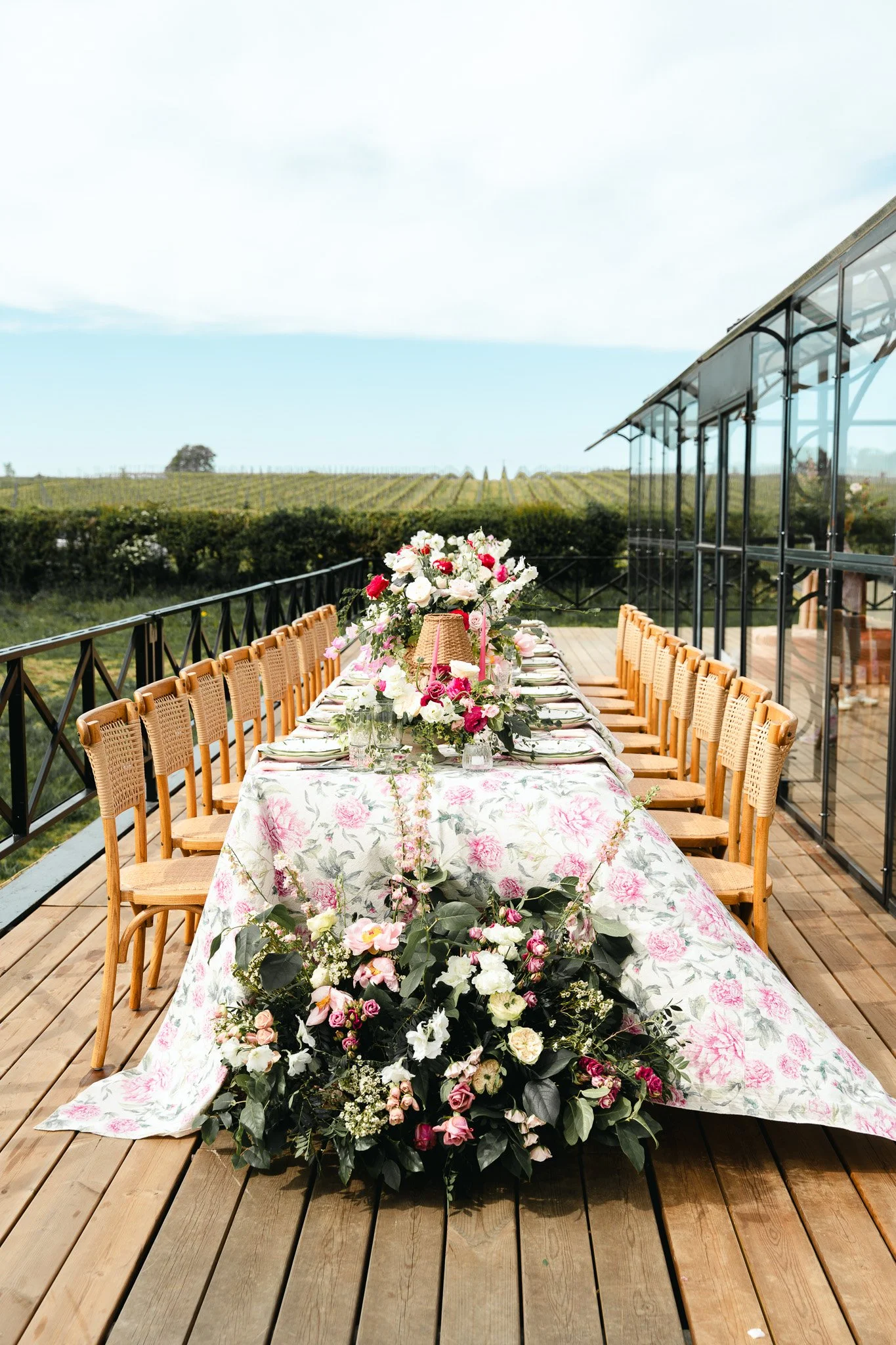 An outdoor dining table decorated with a floral tablecloth and a large floral centerpiece, set on a wooden deck overlooking a vineyard with lush green rows of grapevines.
