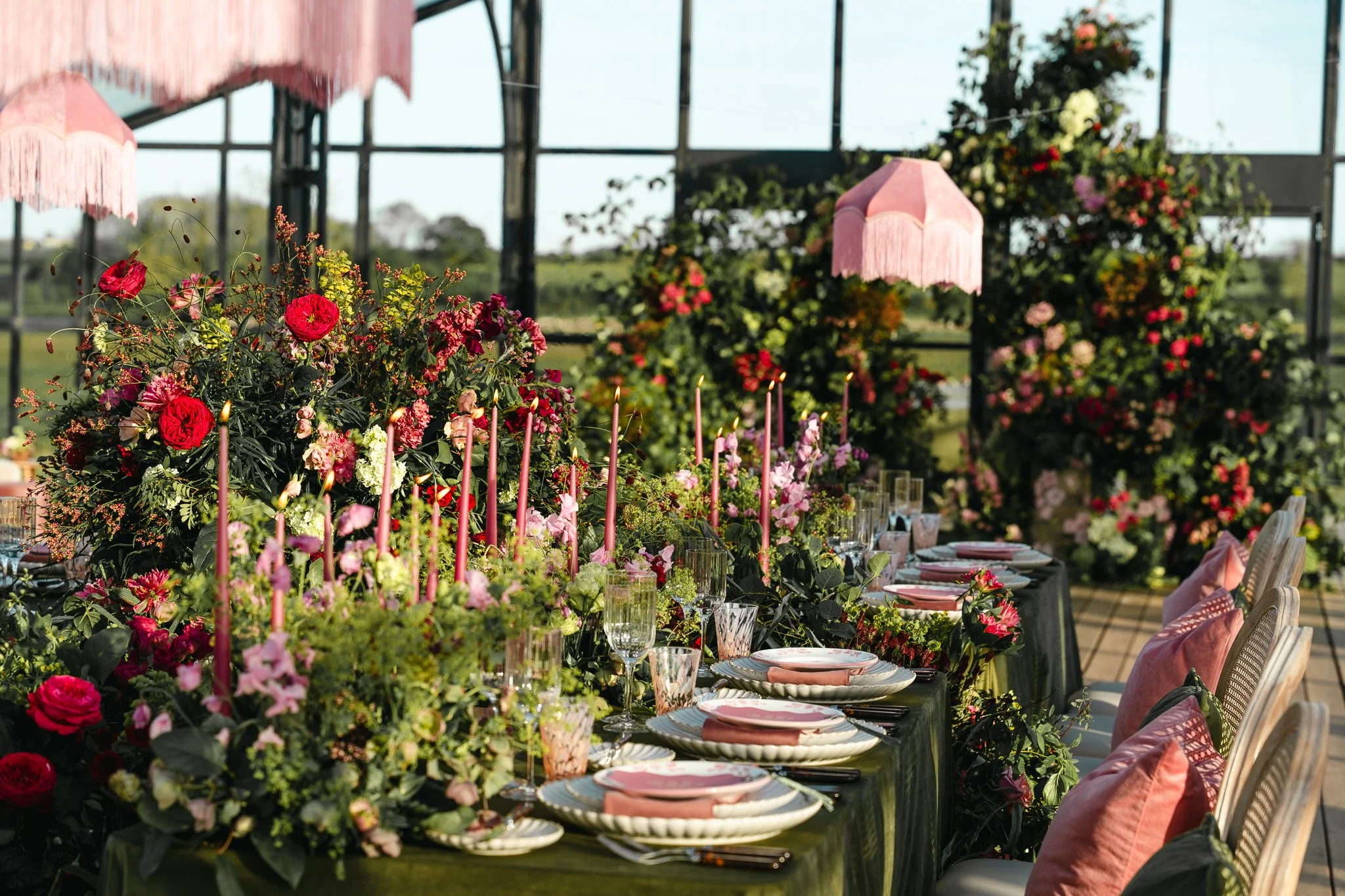A decorated outdoor dining table with pink and red flowers, candles, and wine glasses, set for an event with pink lampshades hanging above, and greenery in the background.