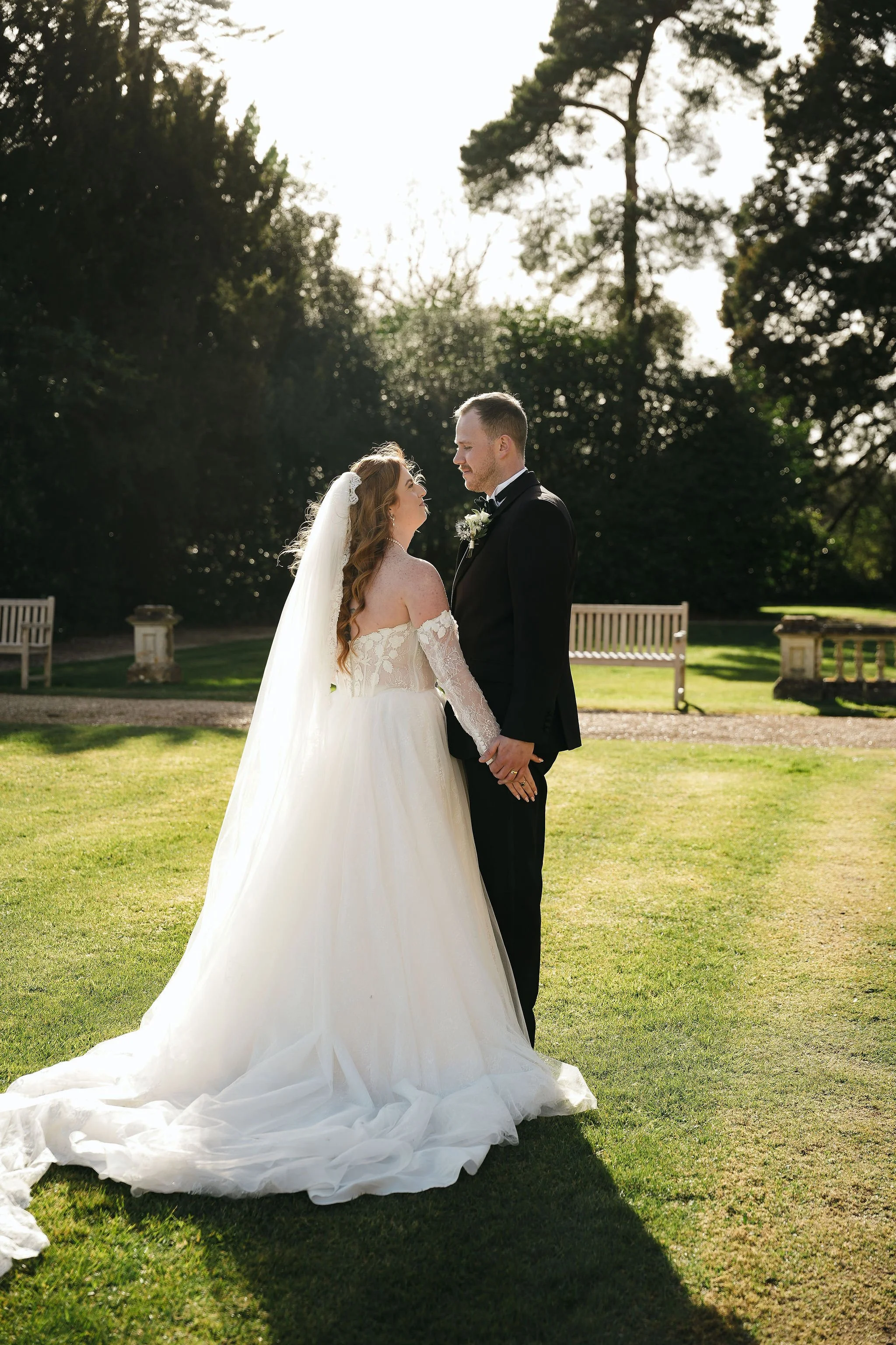 A bride and groom standing outdoors on a grassy area with trees in the background. The bride is wearing a long white wedding gown with lace sleeves and a veil, and the groom is dressed in a black tuxedo with a bow tie. They are facing each other, hol
