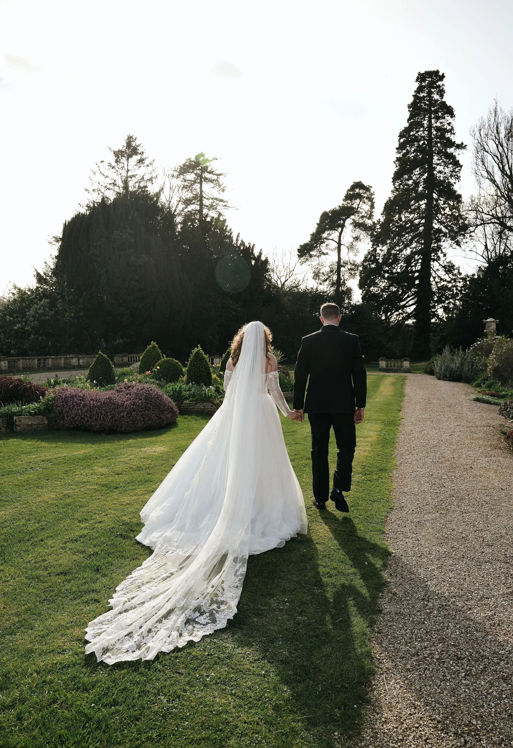 A bride and groom walking hand in hand on a garden path during their wedding in a landscaped garden with tall trees and flowering bushes.