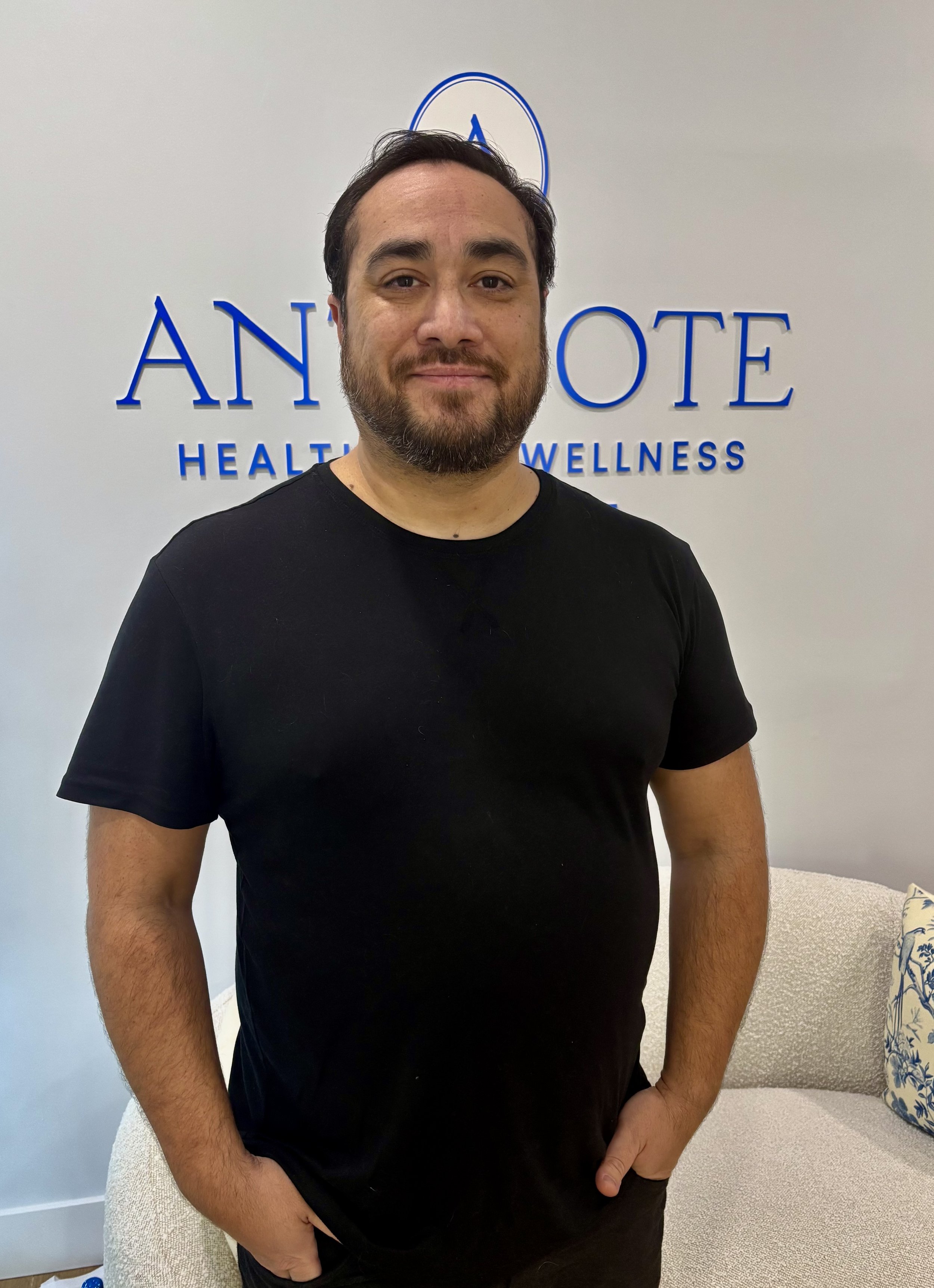 A man with dark hair and a beard wearing a black T-shirt standing inside a wellness center called 'Annote Health & Wellness', with a white wall and a light-colored couch in the background.