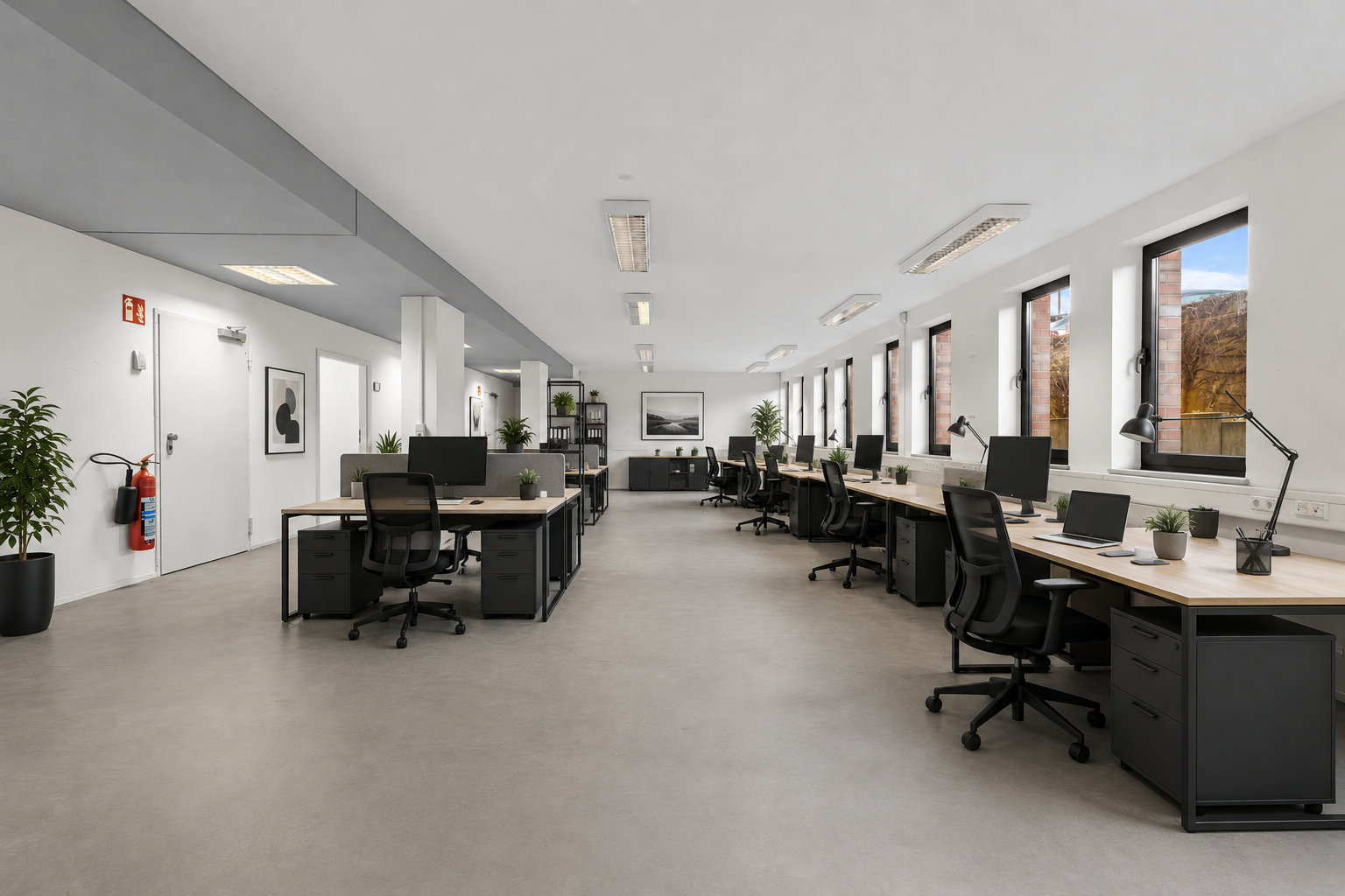 Ein Büro mit mehreren Schreibtischen, schwarzen Bürostühlen, Computerbildschirmen, Pflanzen und großen Fenstern, die Tageslicht hereinlassen.
