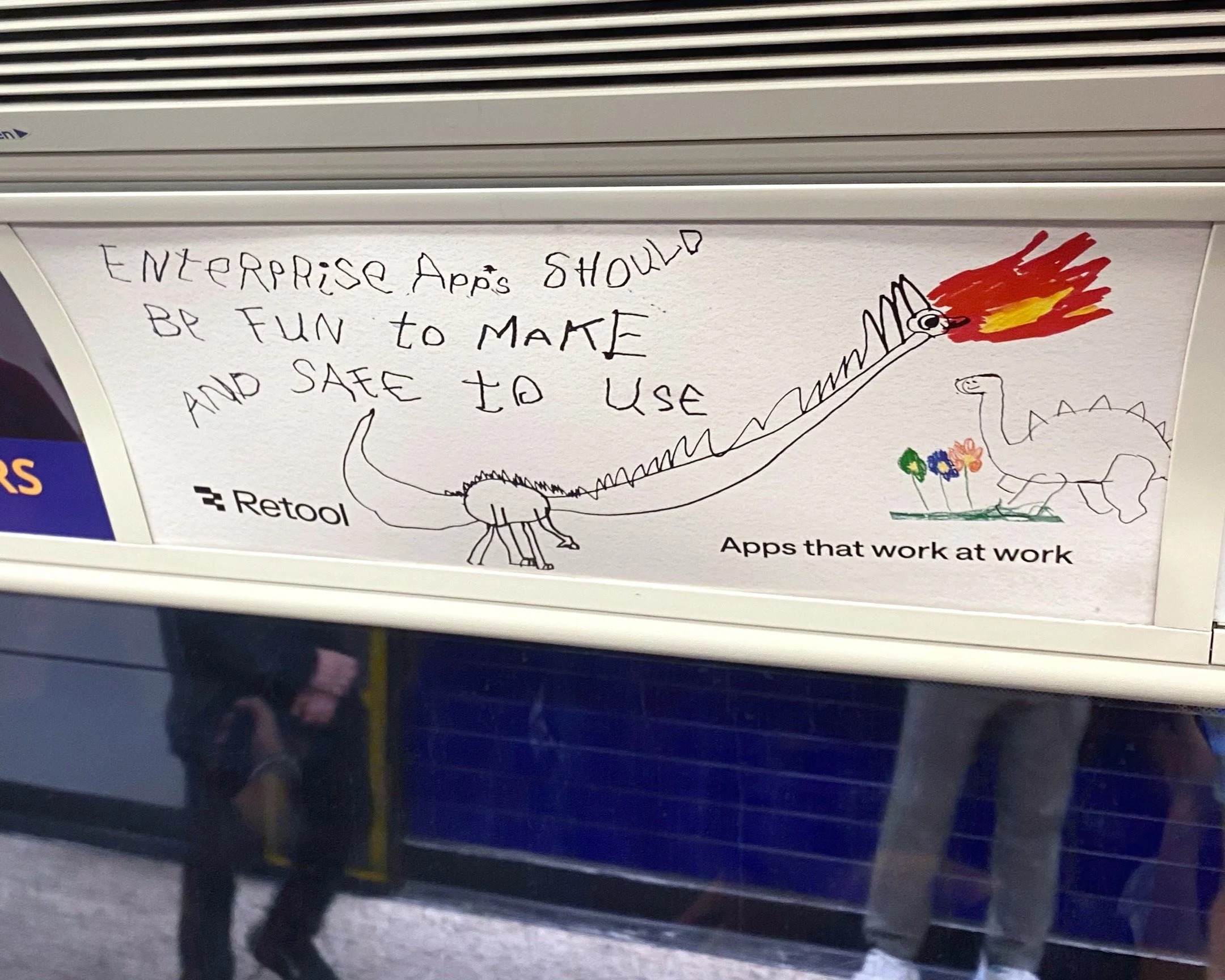 Photo of an advertisement poster above a window in a train, with hand drawn dinosaurs in an infantile style. One them breaths fire in red and yellow crayon.  The poster's slogan is: "Enterprise Apps should be fun to make and safe to use."