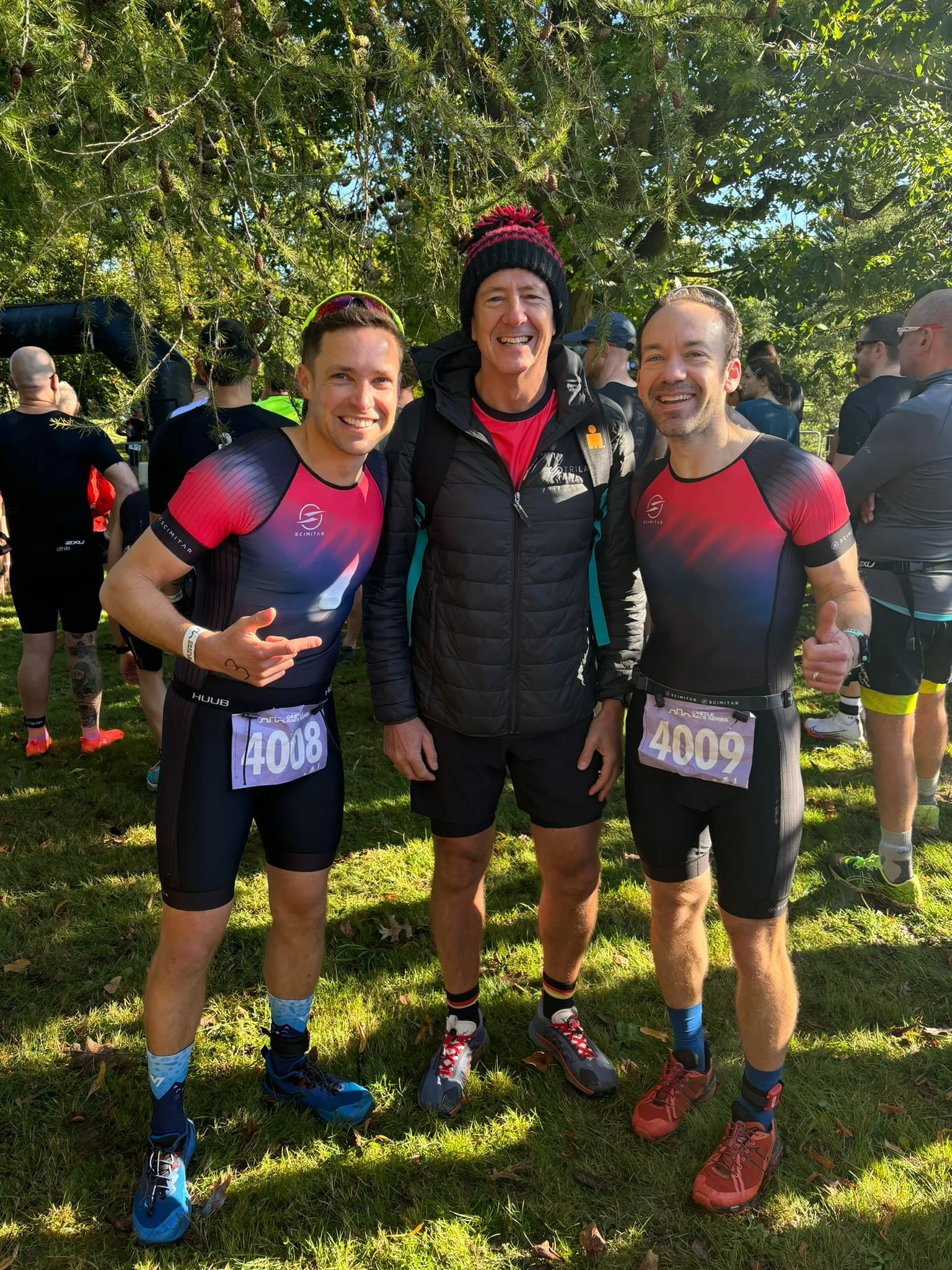Three men posing for a photo at an outdoor event, wearing running gear with race bibs, with trees in the background and other participants gathered around.