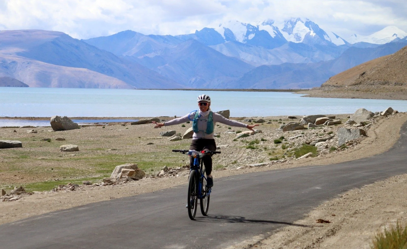 Person riding a bicycle on a paved road along a lake with mountains in the background.