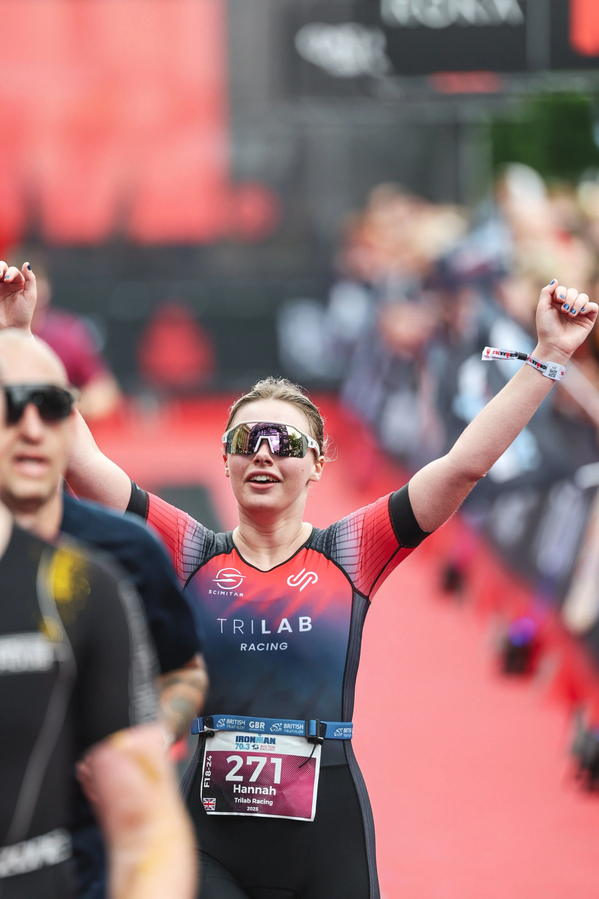 A female athlete wearing sports gear and sunglasses, raising her arms in celebration during a race. She has a race bib reading 'Hannah, 221, Trílab Racing,' and is in a crowded race environment.