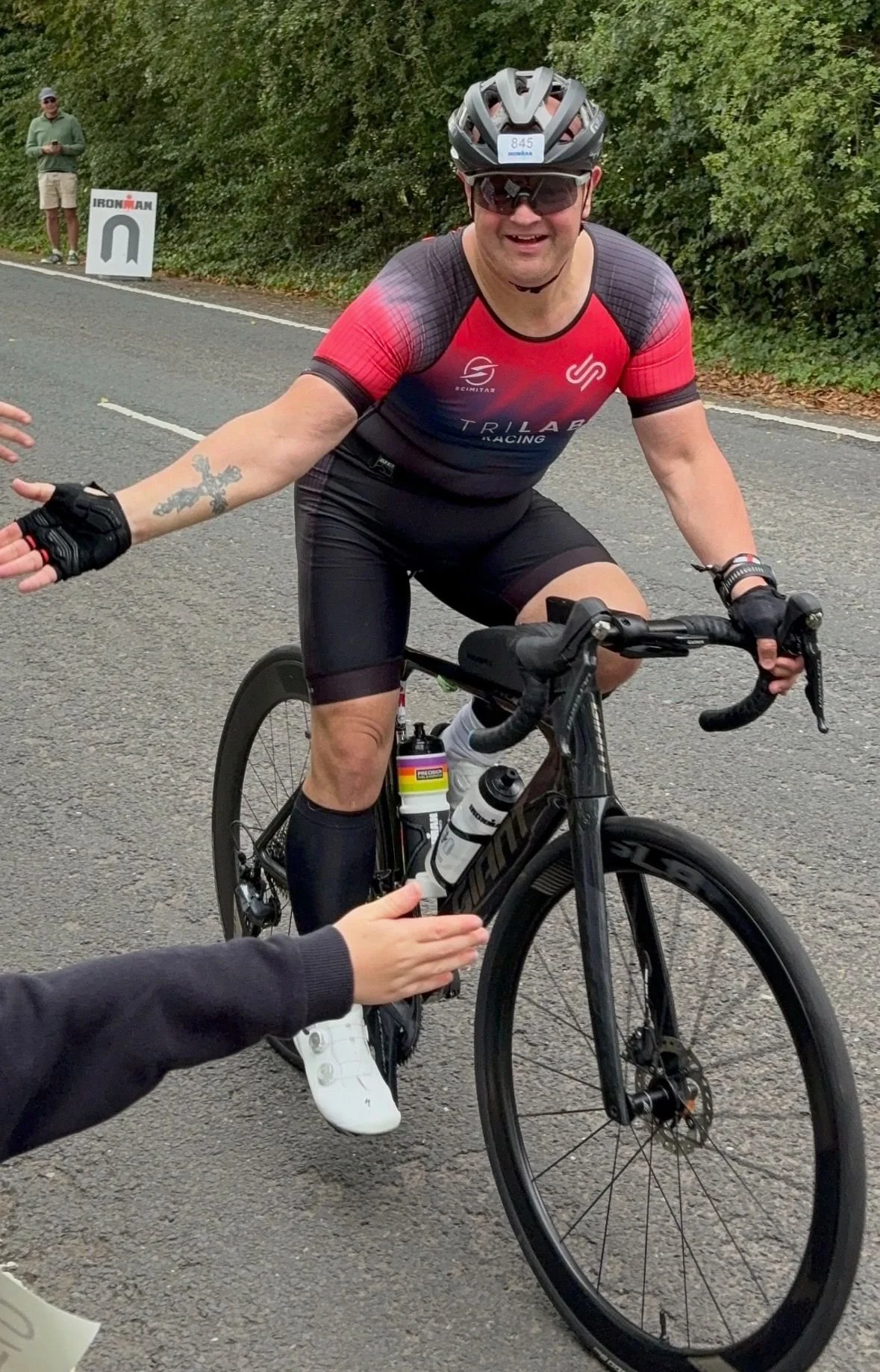 A smiling man wearing a red and black triathlon racing shirt, black cycling shorts, a helmet, and sunglasses, riding a black Trek road bike on a paved road during a race. A volunteer or spectator is handing him a water bottle, and a person in the bac