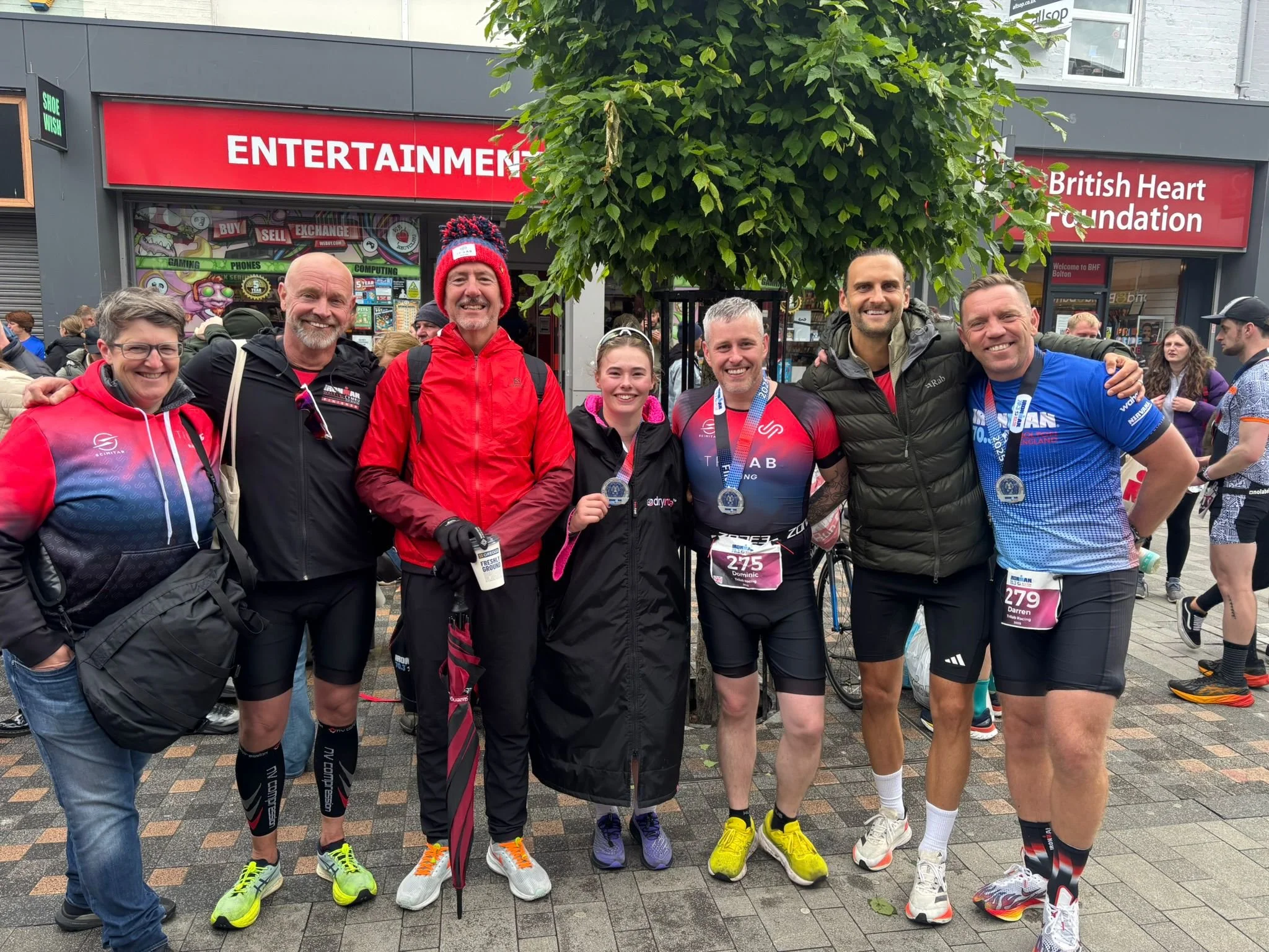 Group of seven smiling marathon runners standing in front of a tree and a building, wearing athletic clothing and medals, with some holding their race bibs.