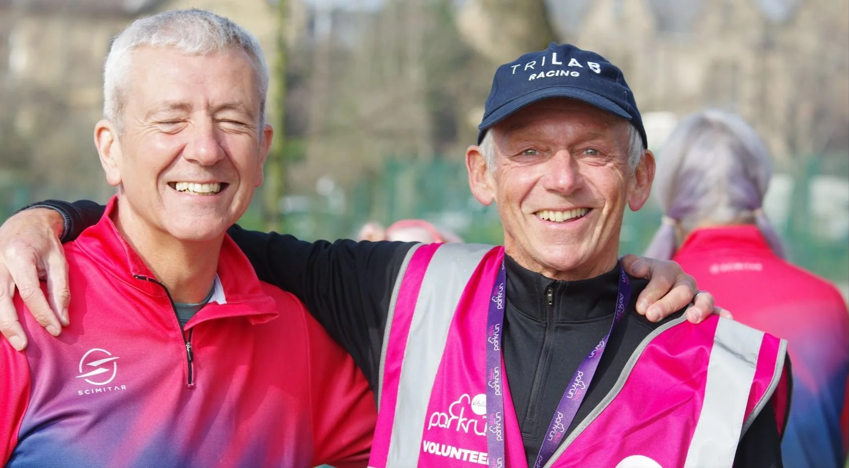 Two smiling men with gray hair and light skin, one wearing a pink and purple sports jacket with a logo and the other wearing a pink volunteer vest, are standing outdoors with their arms around each other, celebrating after a race or event.