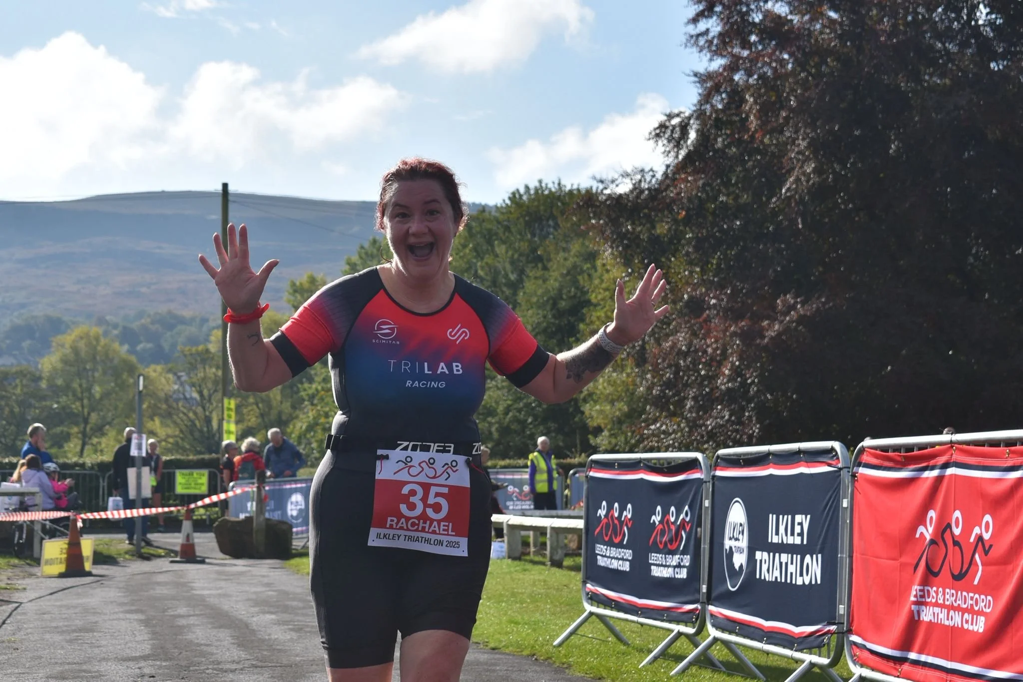 Woman celebrating at the finish line of a triathlon, wearing a red, blue, and black triathlon suit with race number 35, in Ilkley, England, on a sunny day with trees and mountains in the background.