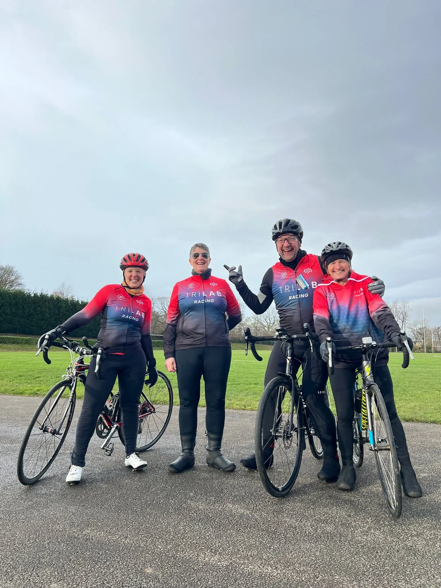 Four people in cycling gear standing with bikes outdoors on a cloudy day, smiling and posing for a photo.