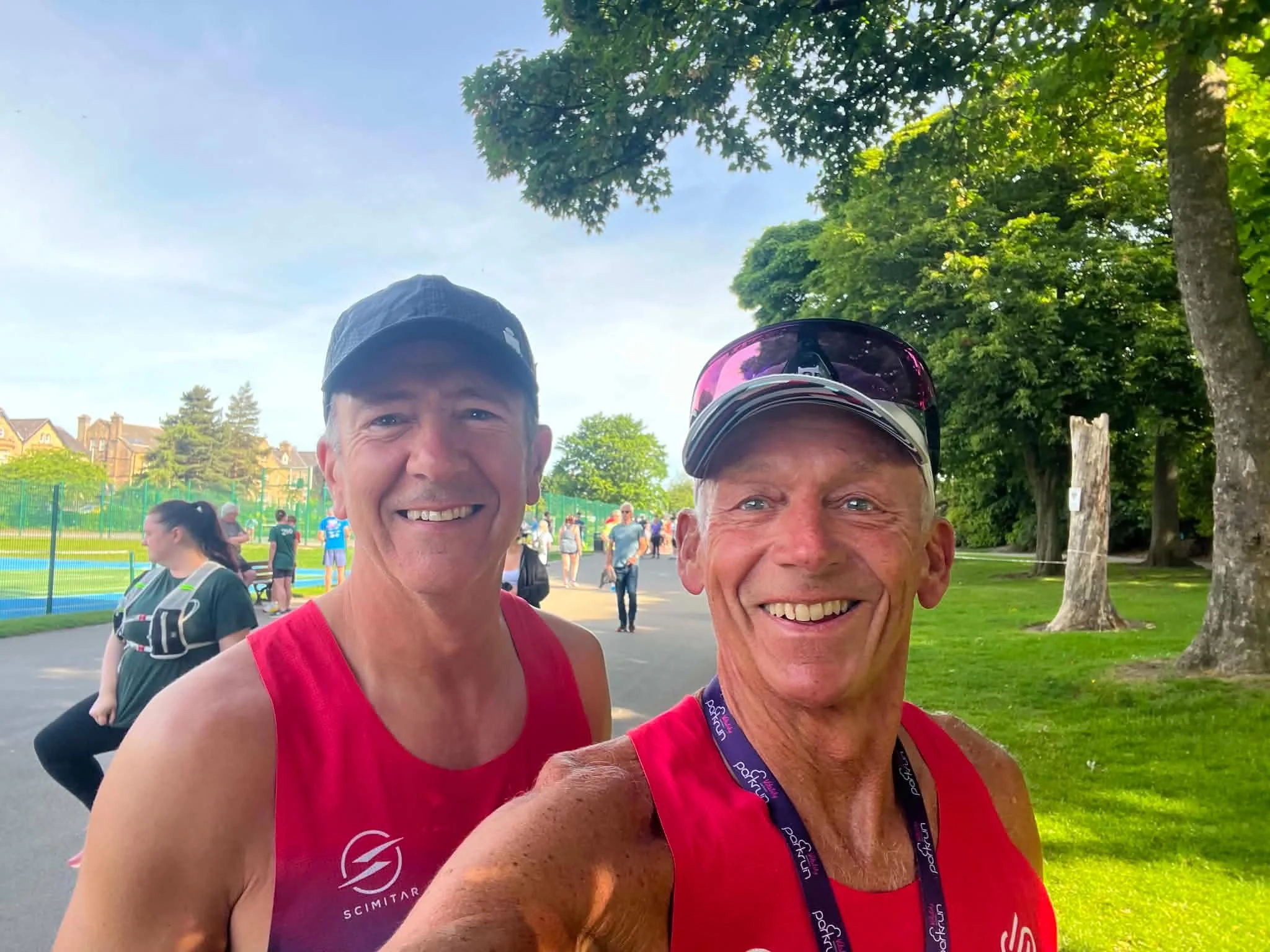 Two smiling men in red athletic shirts taking a selfie at an outdoor event with trees and people in the background.