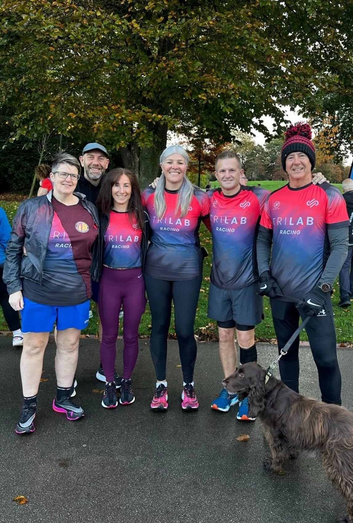 Group of five people in athletic gear, five women and one man, posing outdoors on a cloudy day with green trees in the background. They are smiling and standing on a paved path, with a brown dog on a leash in front of them. Four women are wearing mat