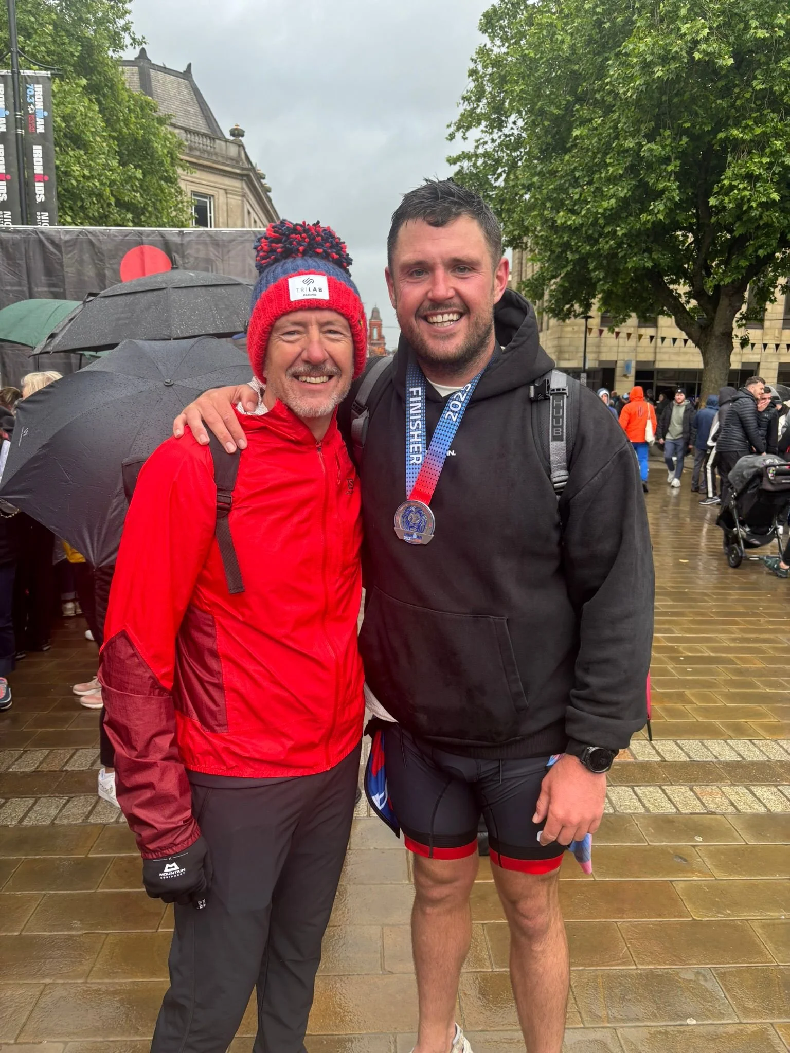 Two men standing together outdoors in a city, smiling at the camera. The man on the left is wearing a red and black jacket, black pants, and a red and blue knit beanie. The man on the right is wearing a black hoodie, black running shorts with red acc