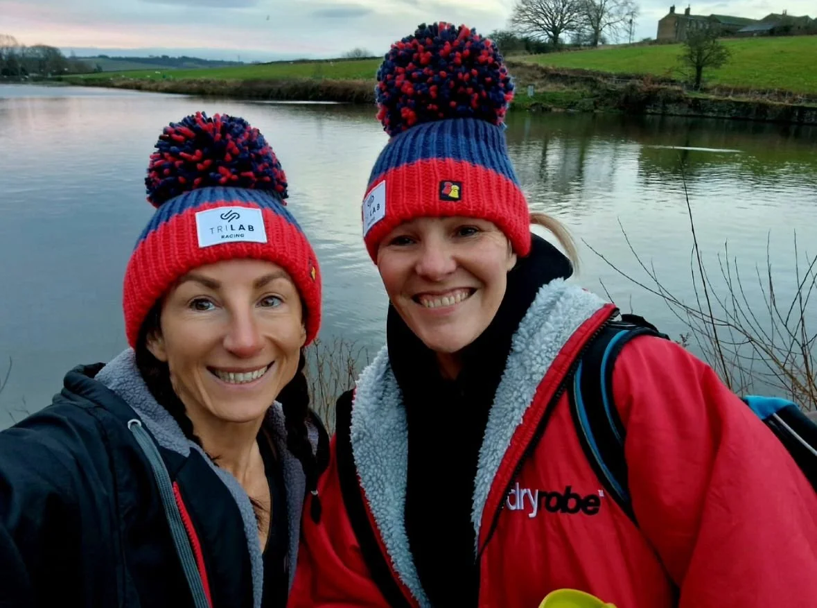 Two women wearing matching red and blue knitted beanies and jackets smiling near a body of water with green fields and trees in the background.